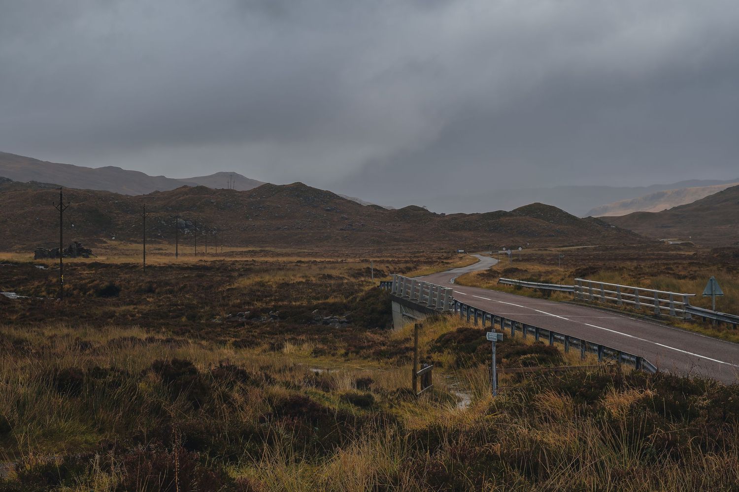 Route goudronnée et pont traversant la lande sauvage des Highlands sous un ciel d'orage menaçant.