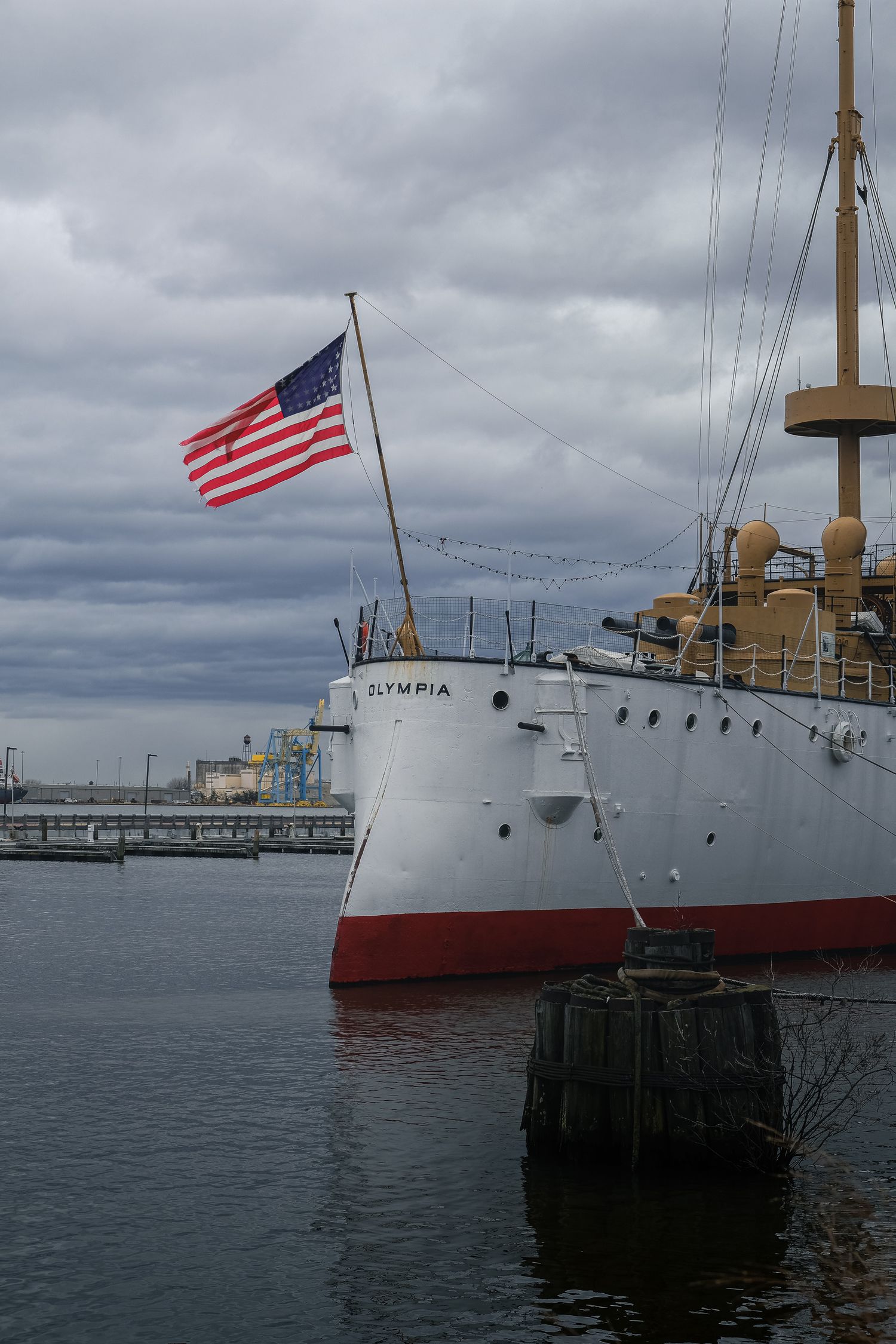 Proue du navire USS Olympia avec drapeau américain à quai, sous un ciel chargé à Philadelphie. Photo de patrimoine naval et historique.