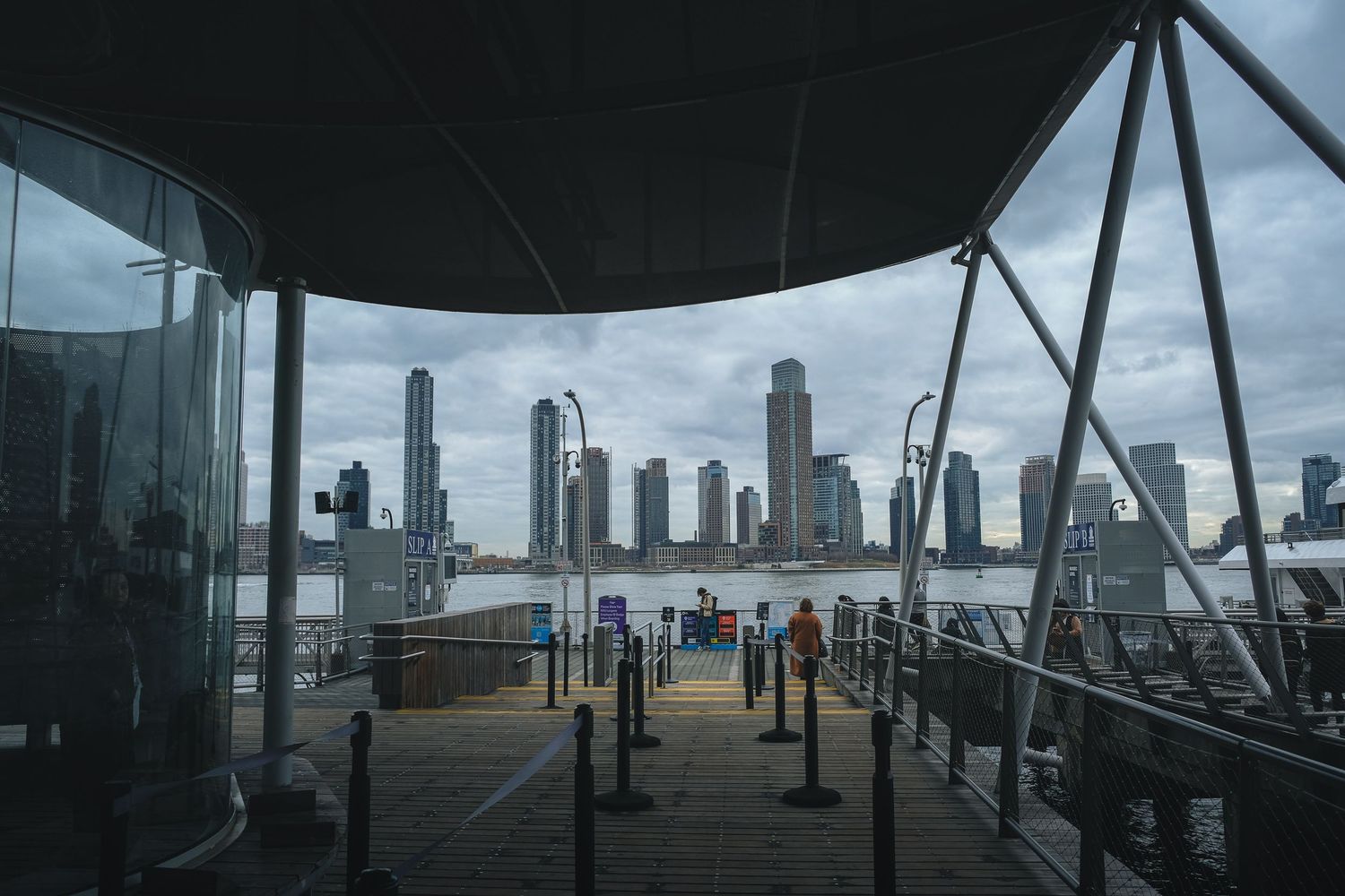 Quai d'embarquement pour ferry à Manhattan face à la skyline de Jersey City sous un ciel couvert. Photo de transport et paysage urbain.