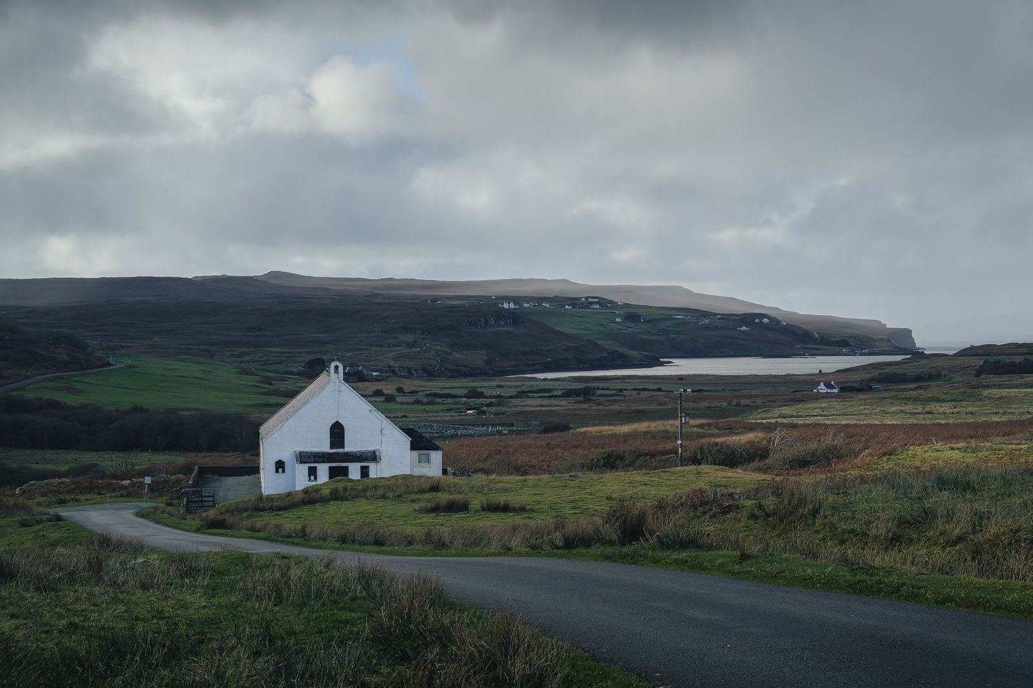 Bâtiment blanc au bord d'une route sinueuse sur l'Île de Skye. Vue sur un loch et les collines d'Écosse sous un ciel nuageux.