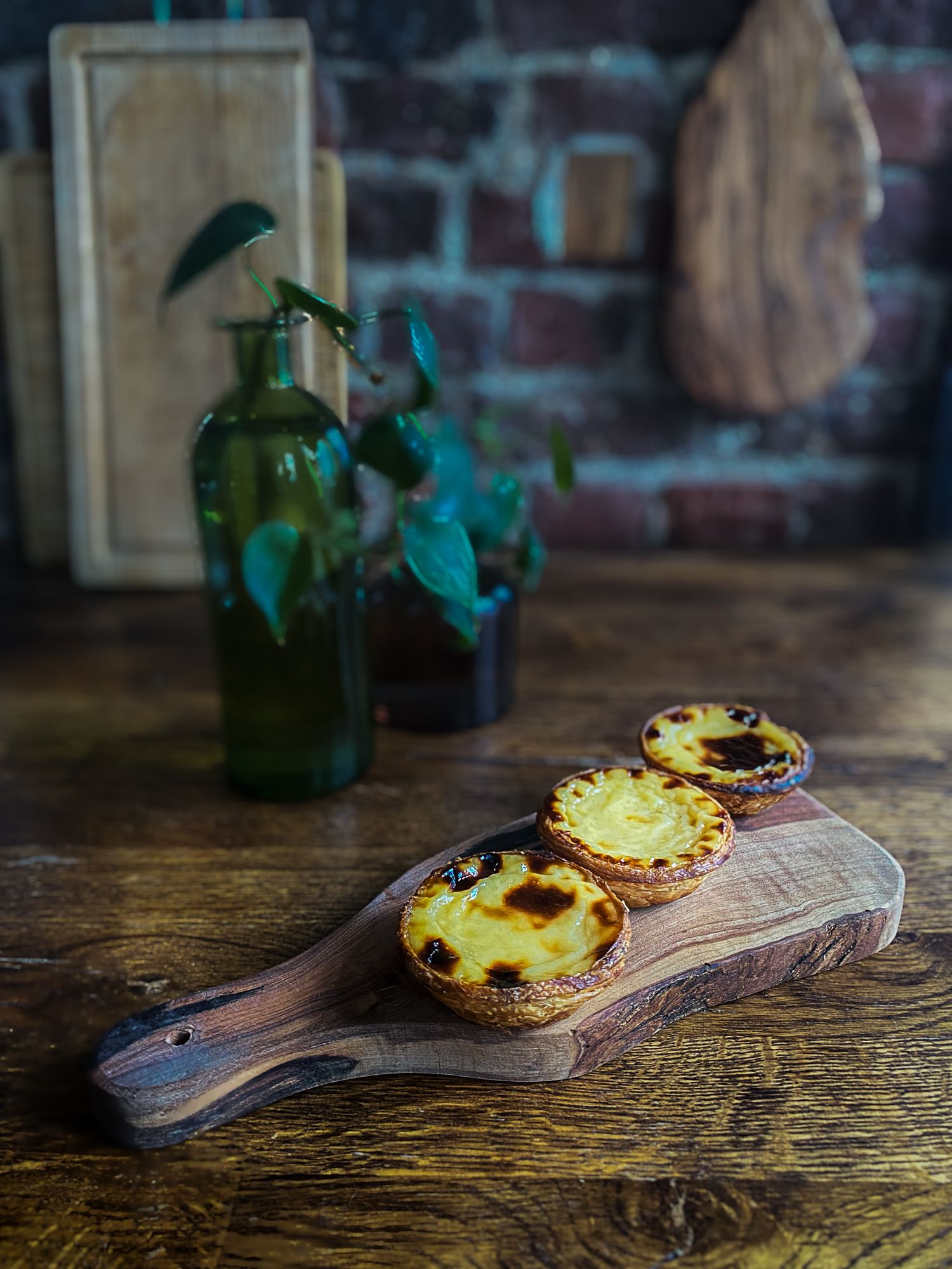 Trois pasteis de nata dorés présentés sur une planche en bois brut, dans une ambiance rustique et chaleureuse. Photographie culinaire.