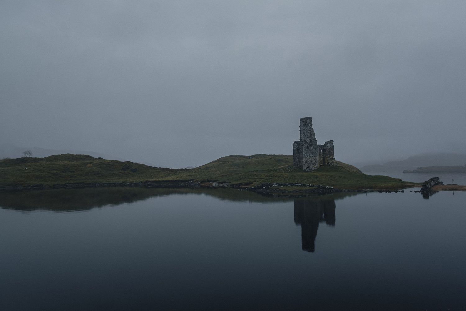 Photographie minimaliste des ruines du château d’Ardvreck en Écosse, reflets sombres sur le Loch Assynt sous un ciel de brume mystique.
