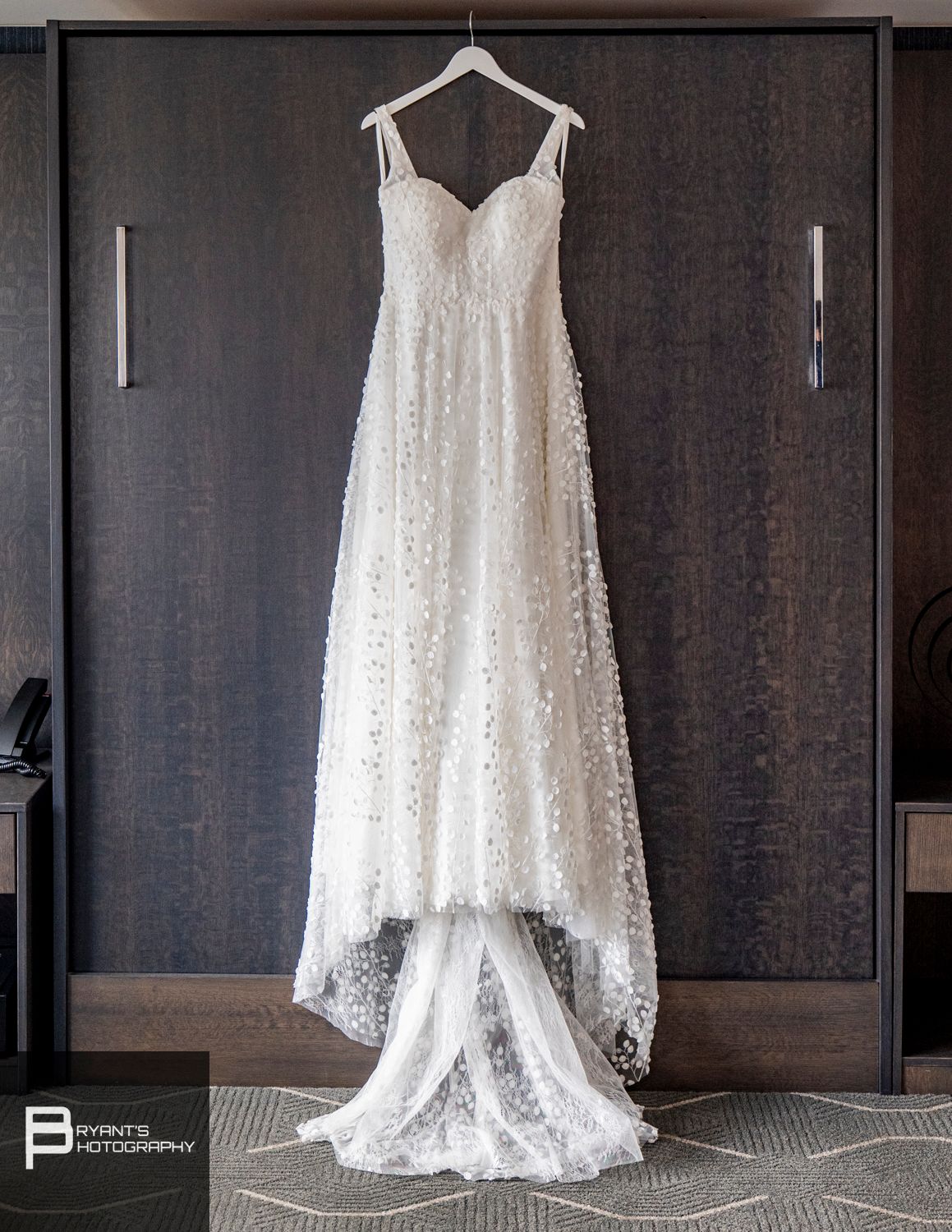 Elegant white lace wedding dress hanging against dark wood paneled wall in hotel room.