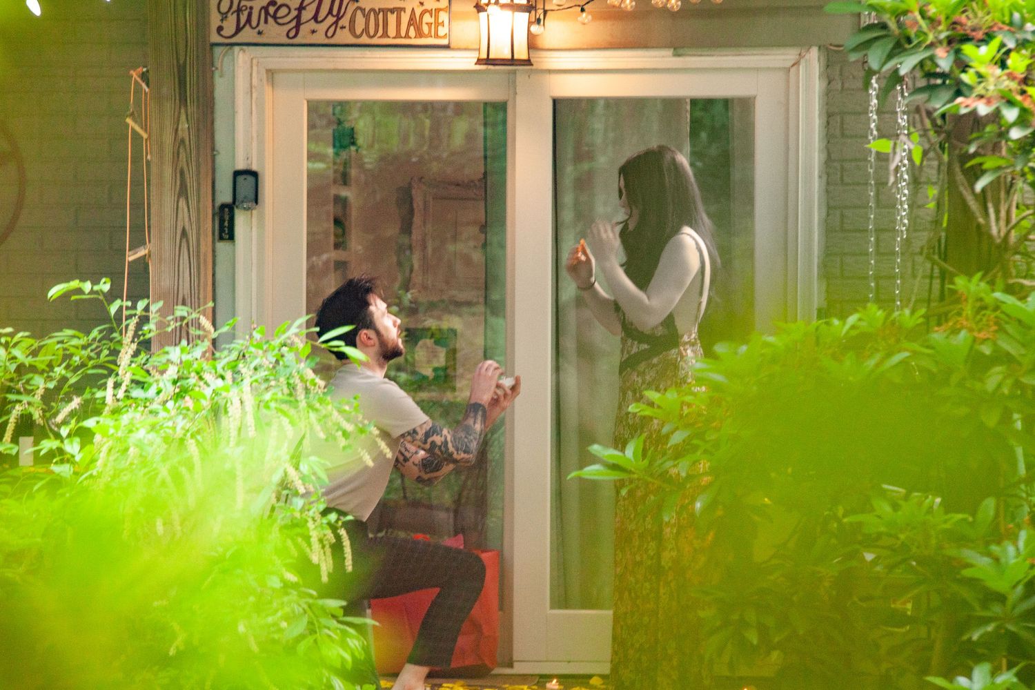 A proposal moment captured through glass doors with string lights and plants.