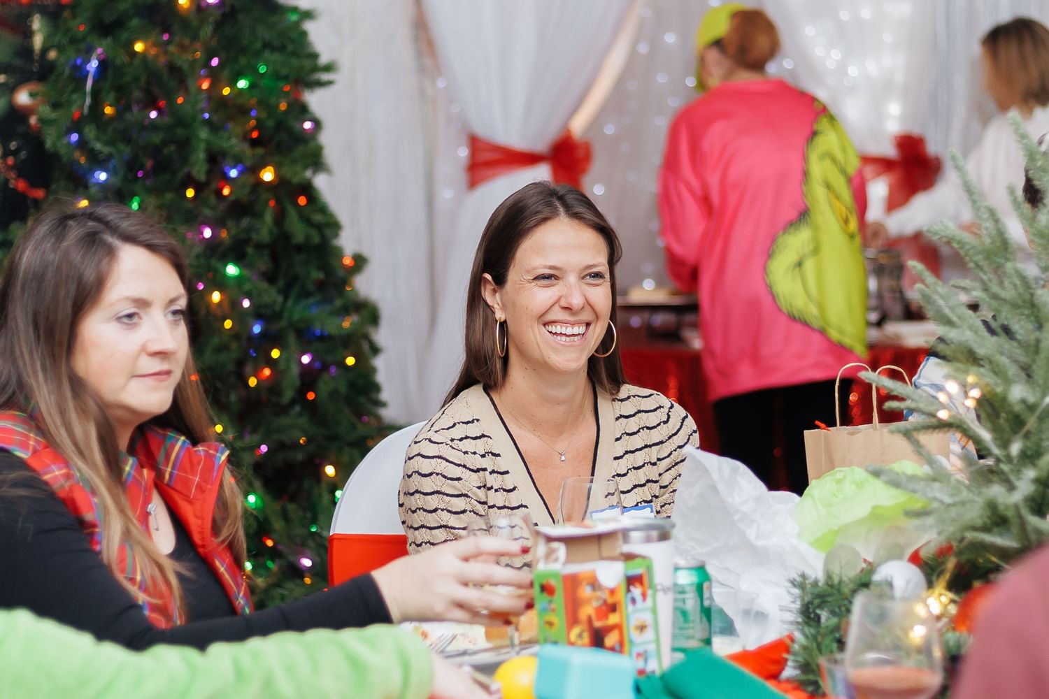 Two women smiling at a festive Christmas party table with gifts, decorations, and a lit Christmas tree in the background.