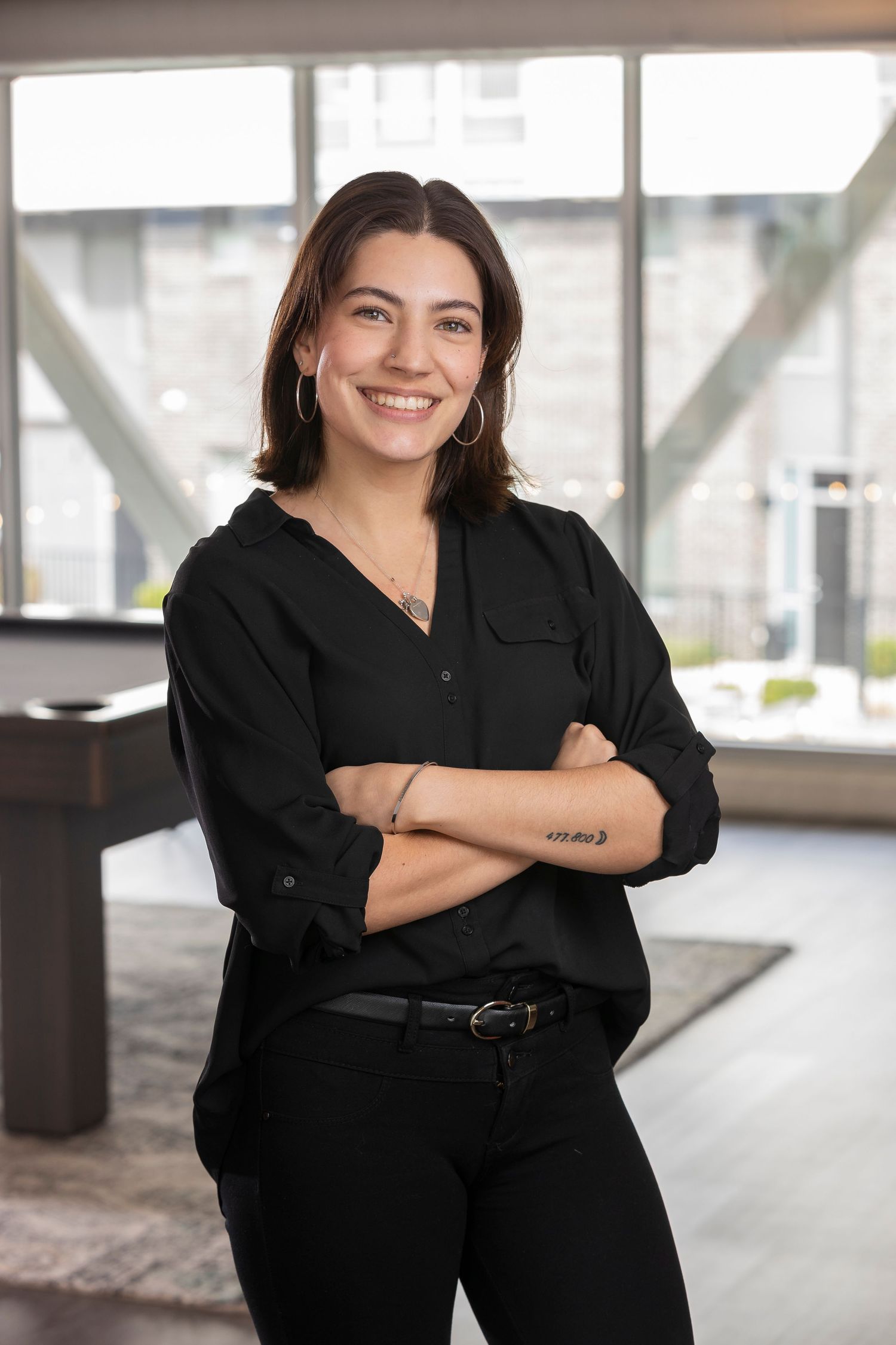 Smiling young woman in black outfit with arms crossed, standing in a modern office space with large windows.
