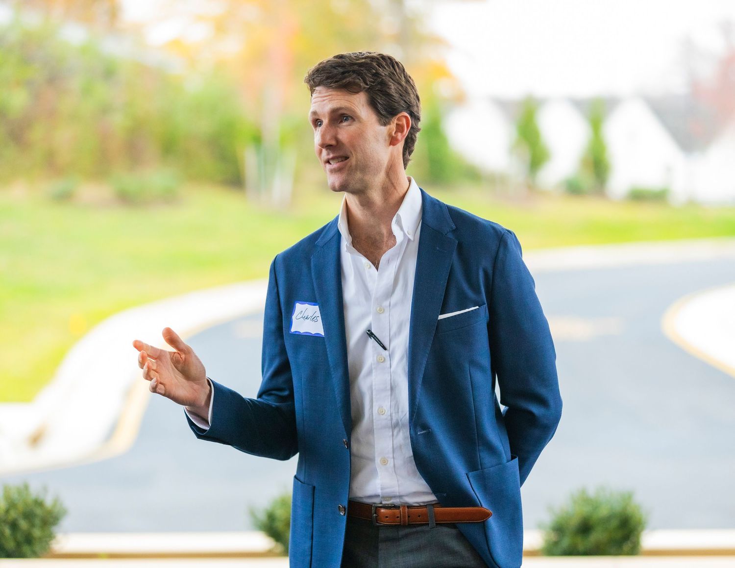 Man in blue blazer speaking outdoors at a professional event, gesturing with hand against green landscape backdrop.