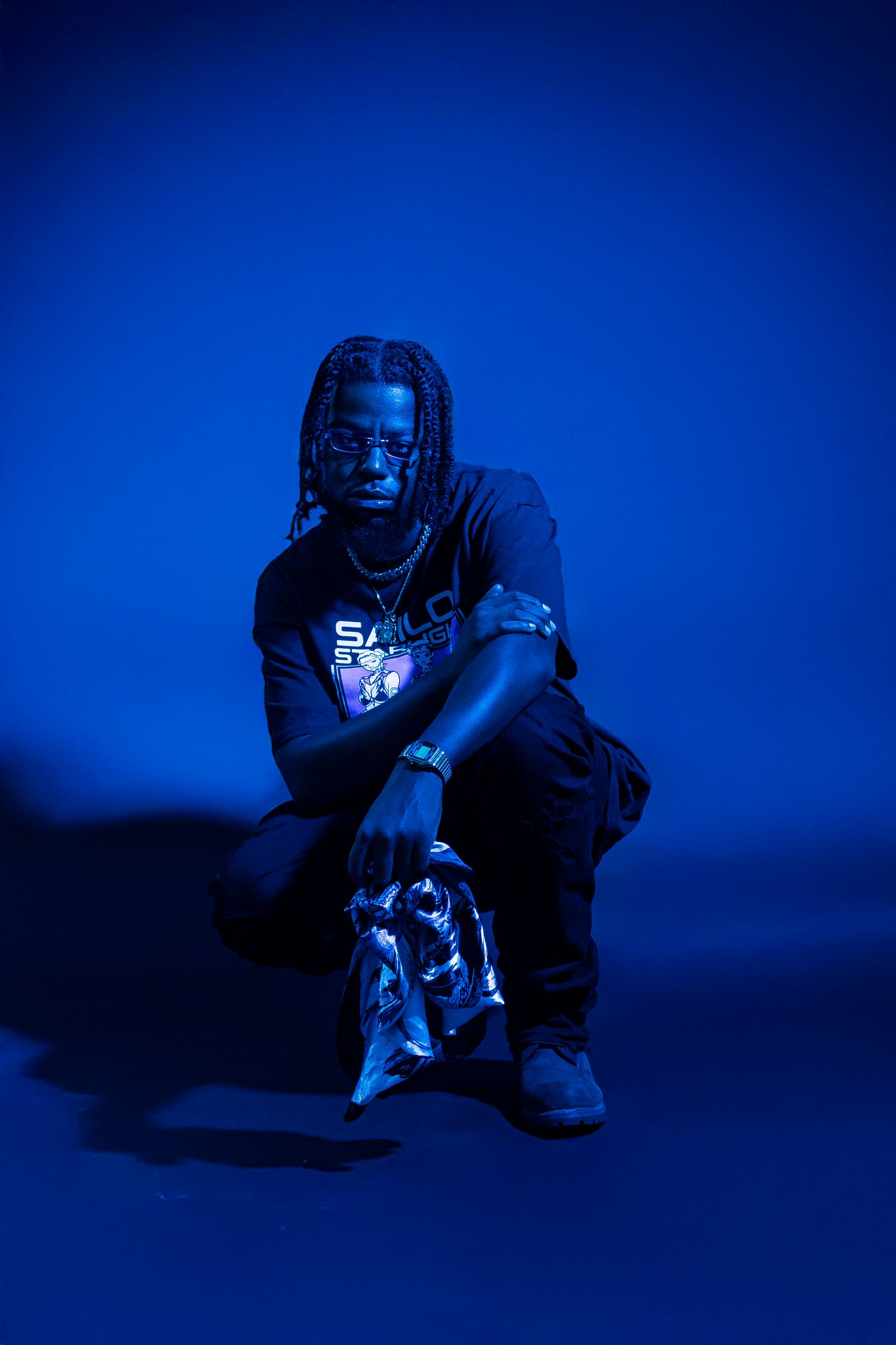 Young man with braids crouching in blue-lit studio, wearing black graphic tee, chains, and dark cargo pants.