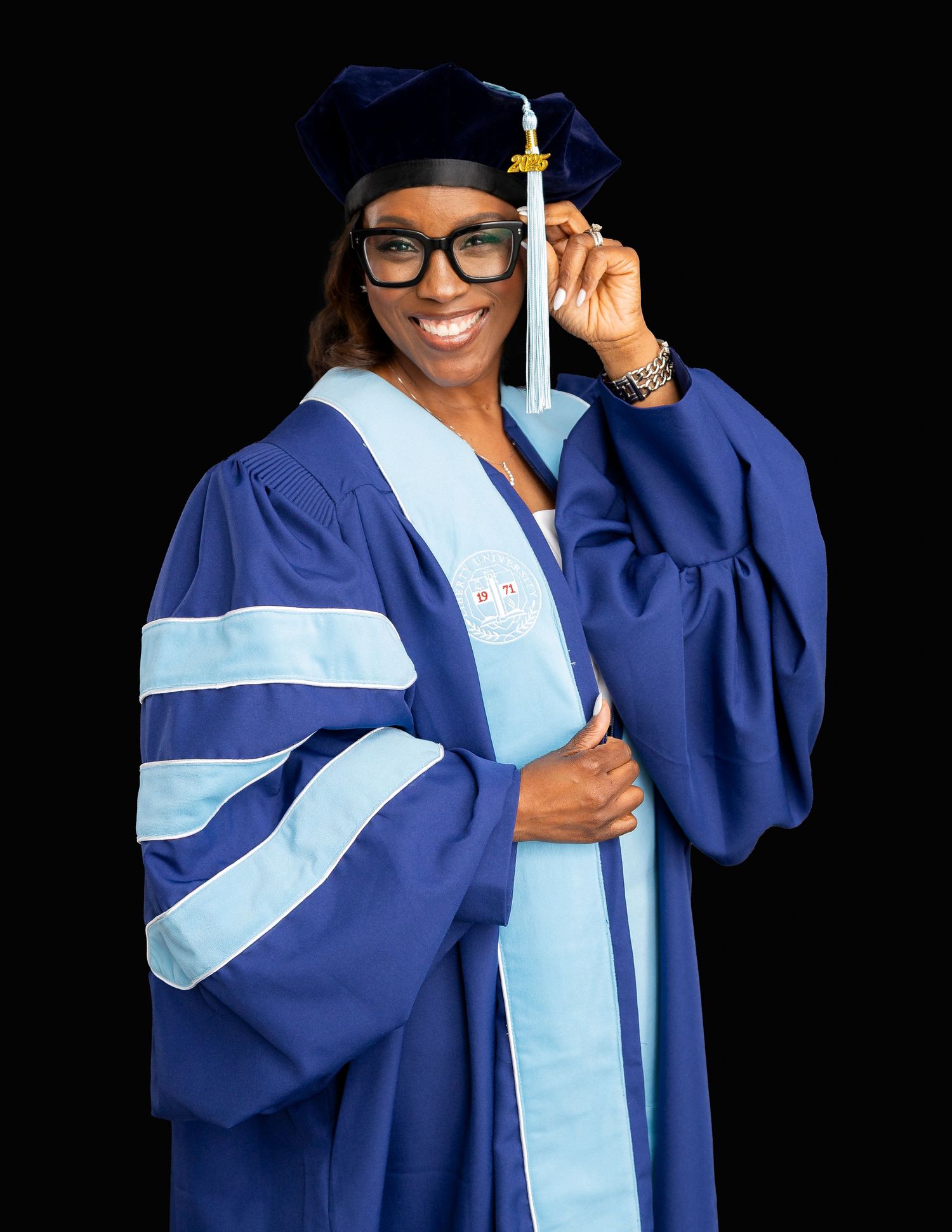 Smiling woman in blue doctoral graduation gown and cap holding tassel against black background.