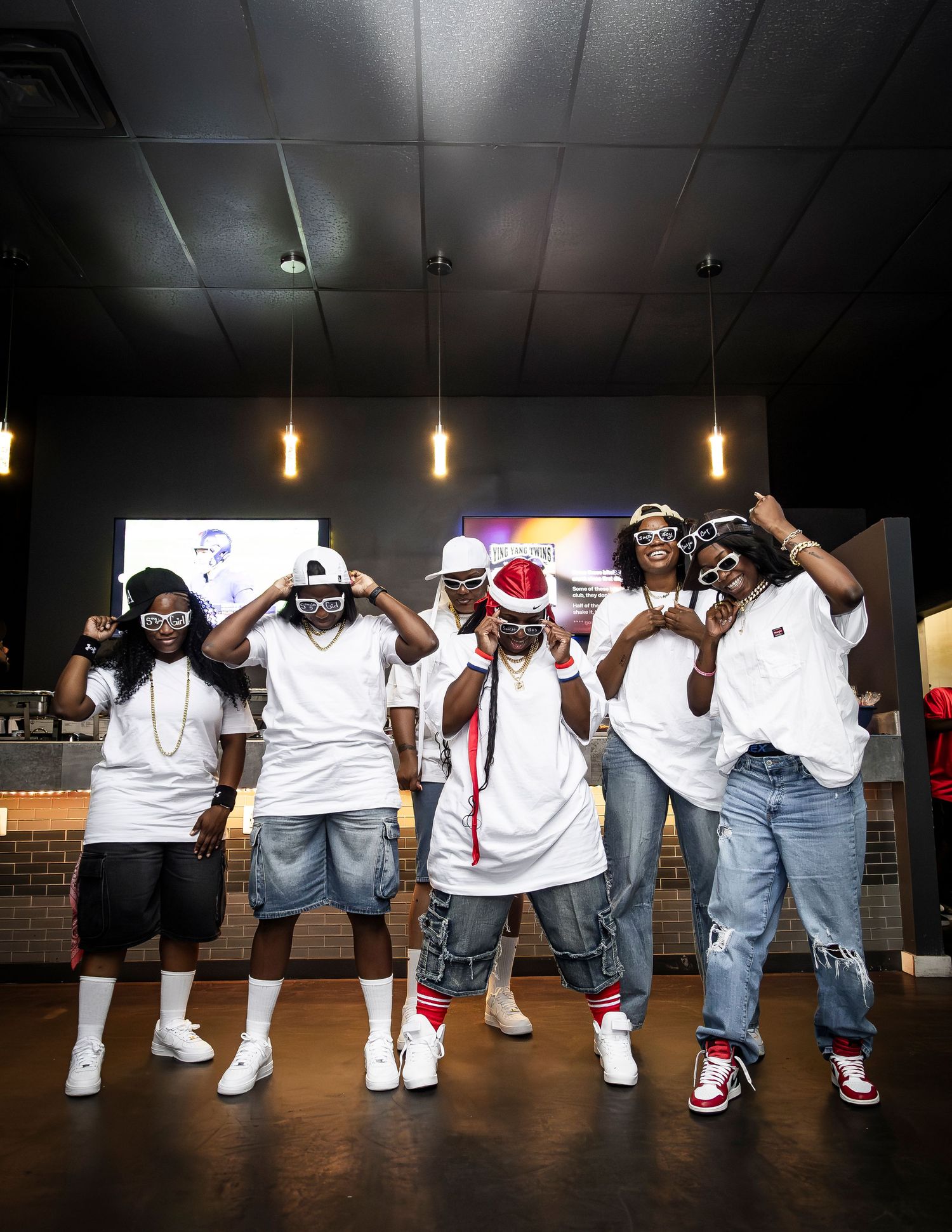 Group of young men in matching white tees posing together inside a modern restaurant with pendant lighting.