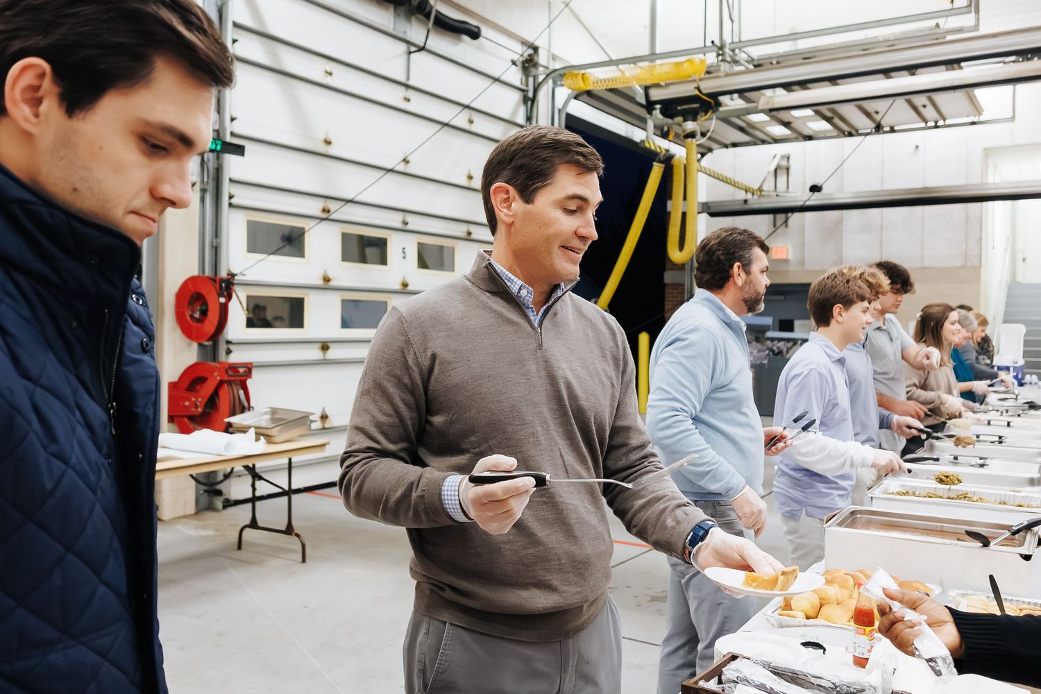 Group of men serving themselves at a buffet line in an industrial workspace during a catered office lunch event.