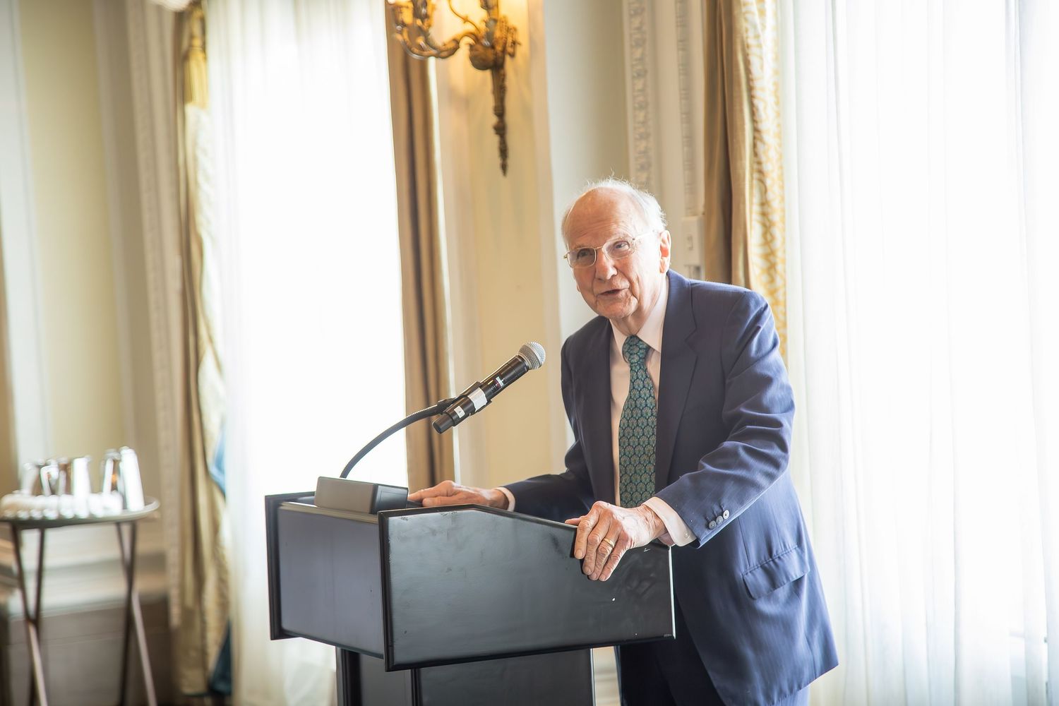 Elderly man in blue suit speaking at a podium in an elegant room with gold curtains and wall sconces.