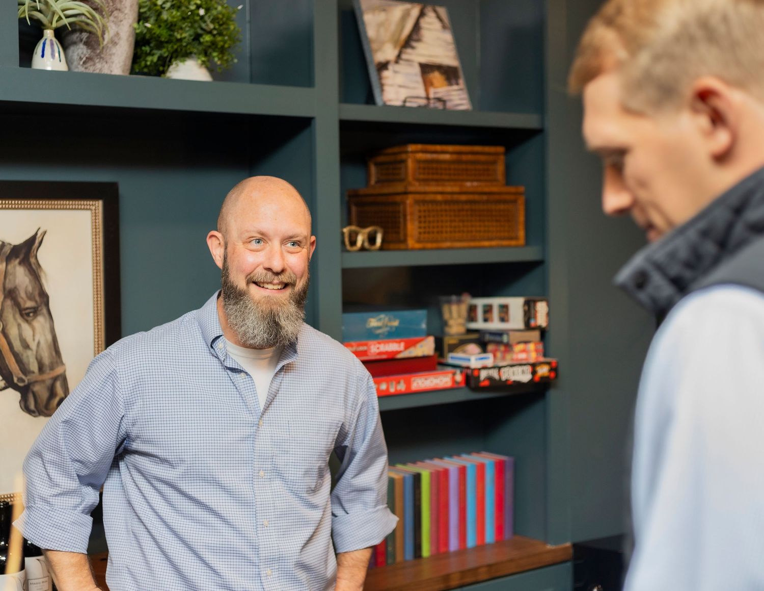 Two men conversing in a stylish home office with dark shelving, books, and a horse painting on the wall.