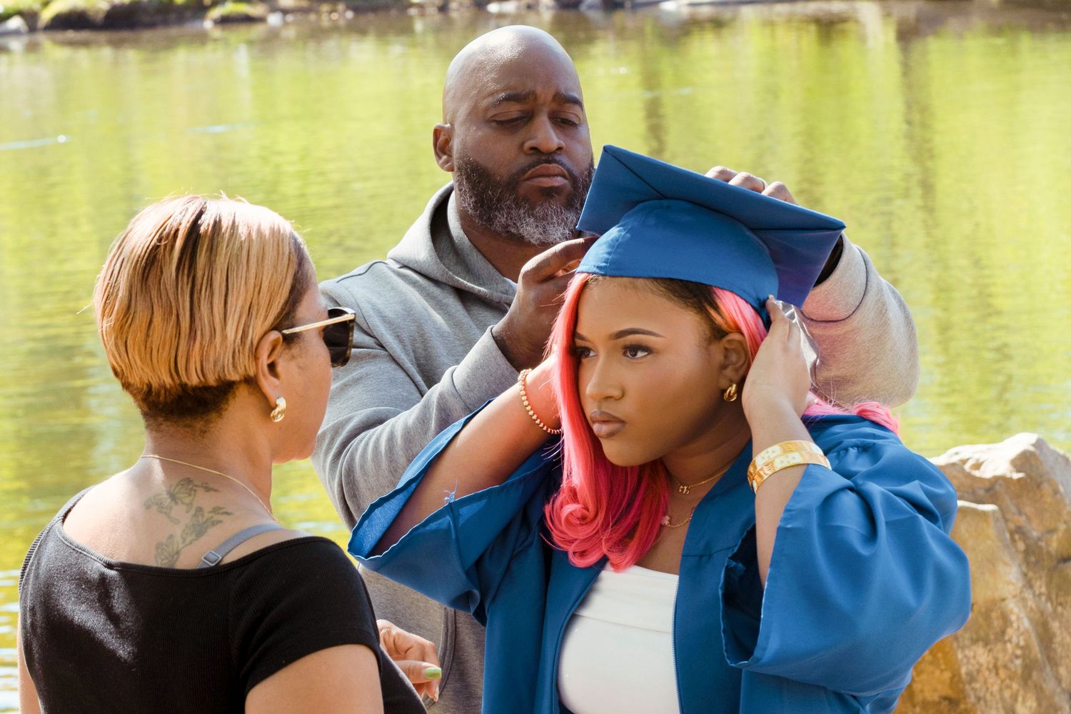 Parents helping their daughter put on a blue graduation cap outdoors near a lake.