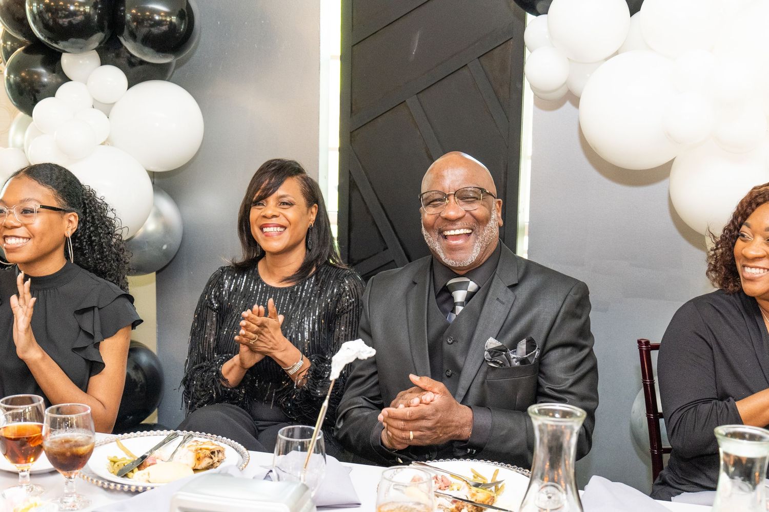 Group of people smiling and clapping at a formal black and white themed event with balloon decorations.
