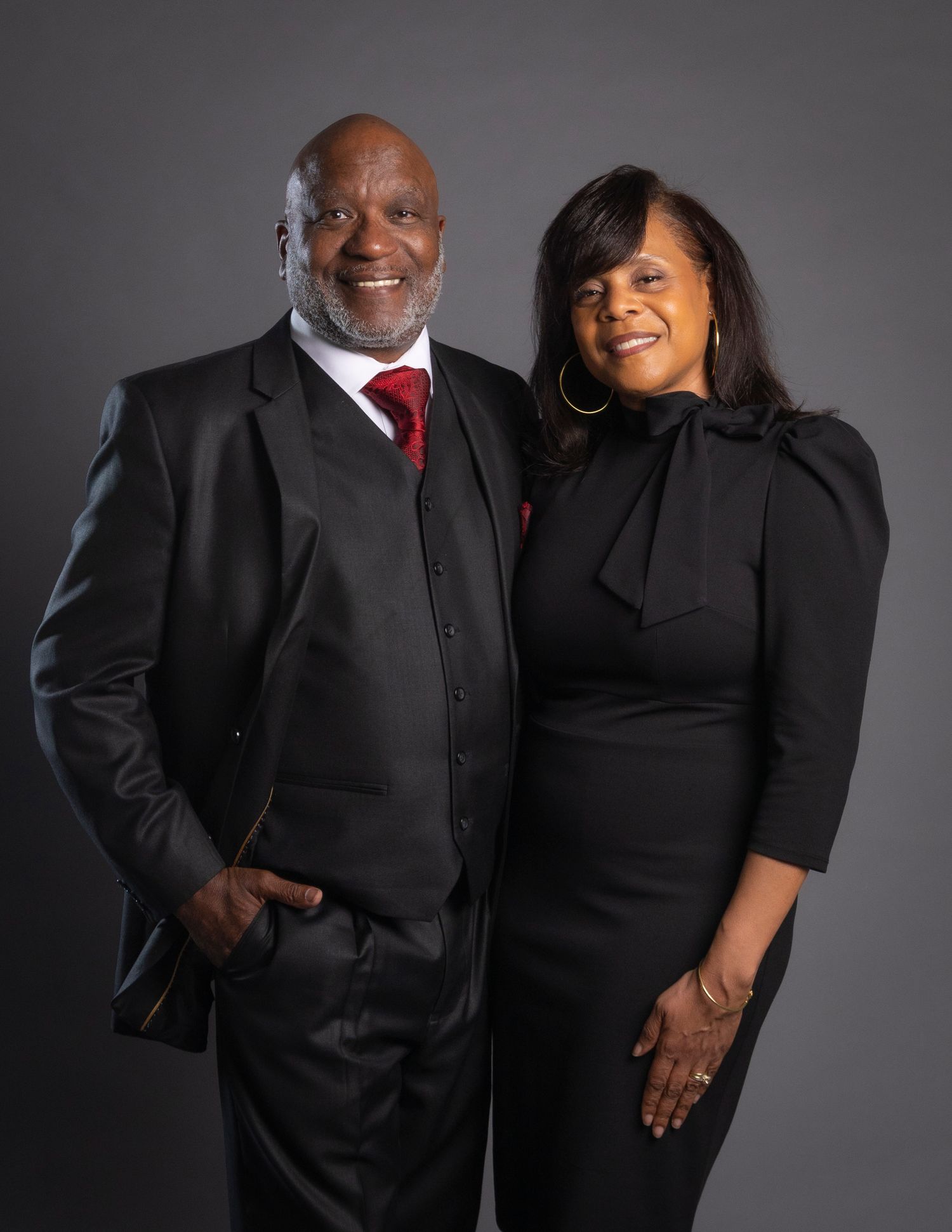 A well-dressed couple in matching black formal attire smiling together against a gray studio background.