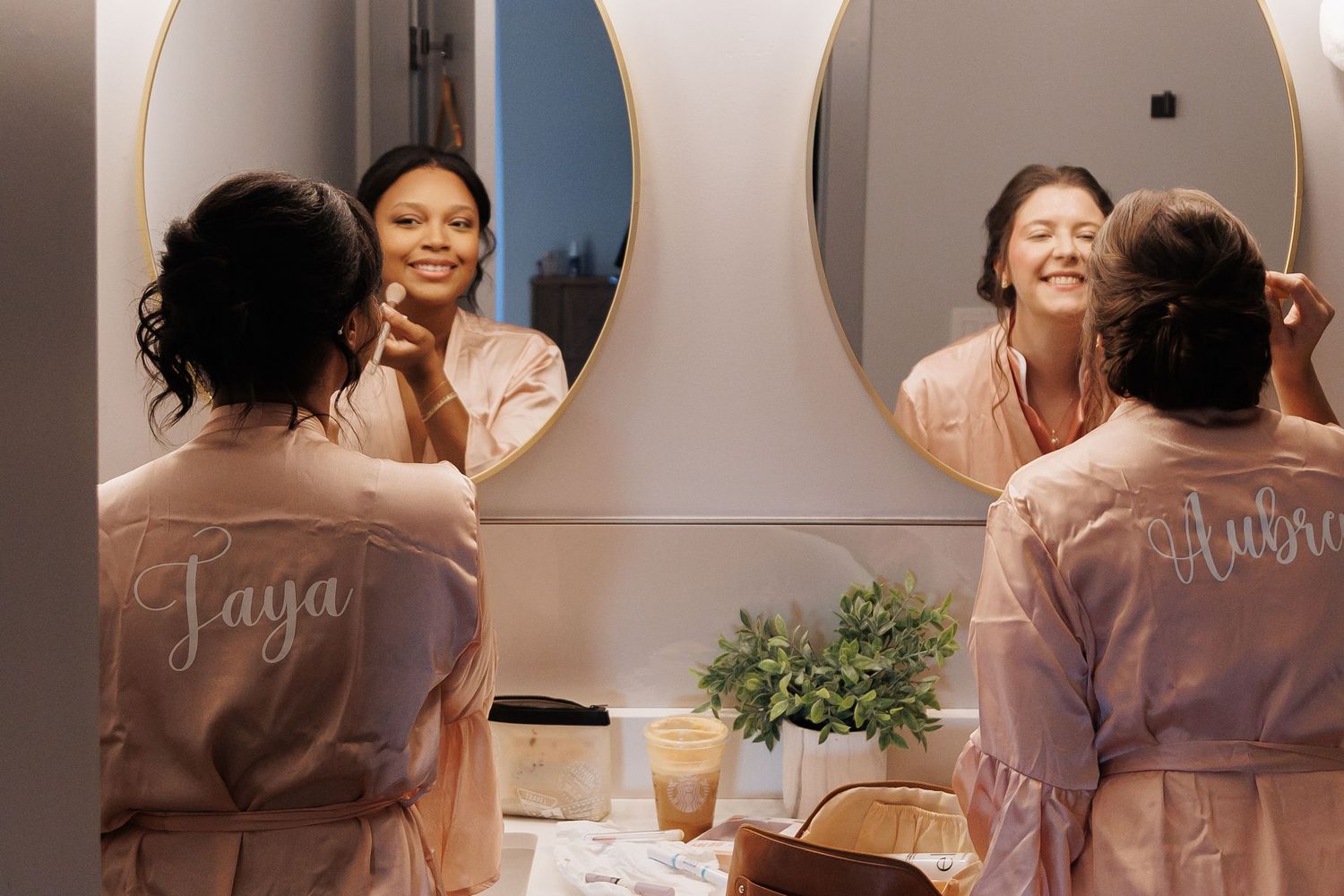 Wedding party getting ready in matching pink robes while looking in mirrors.
