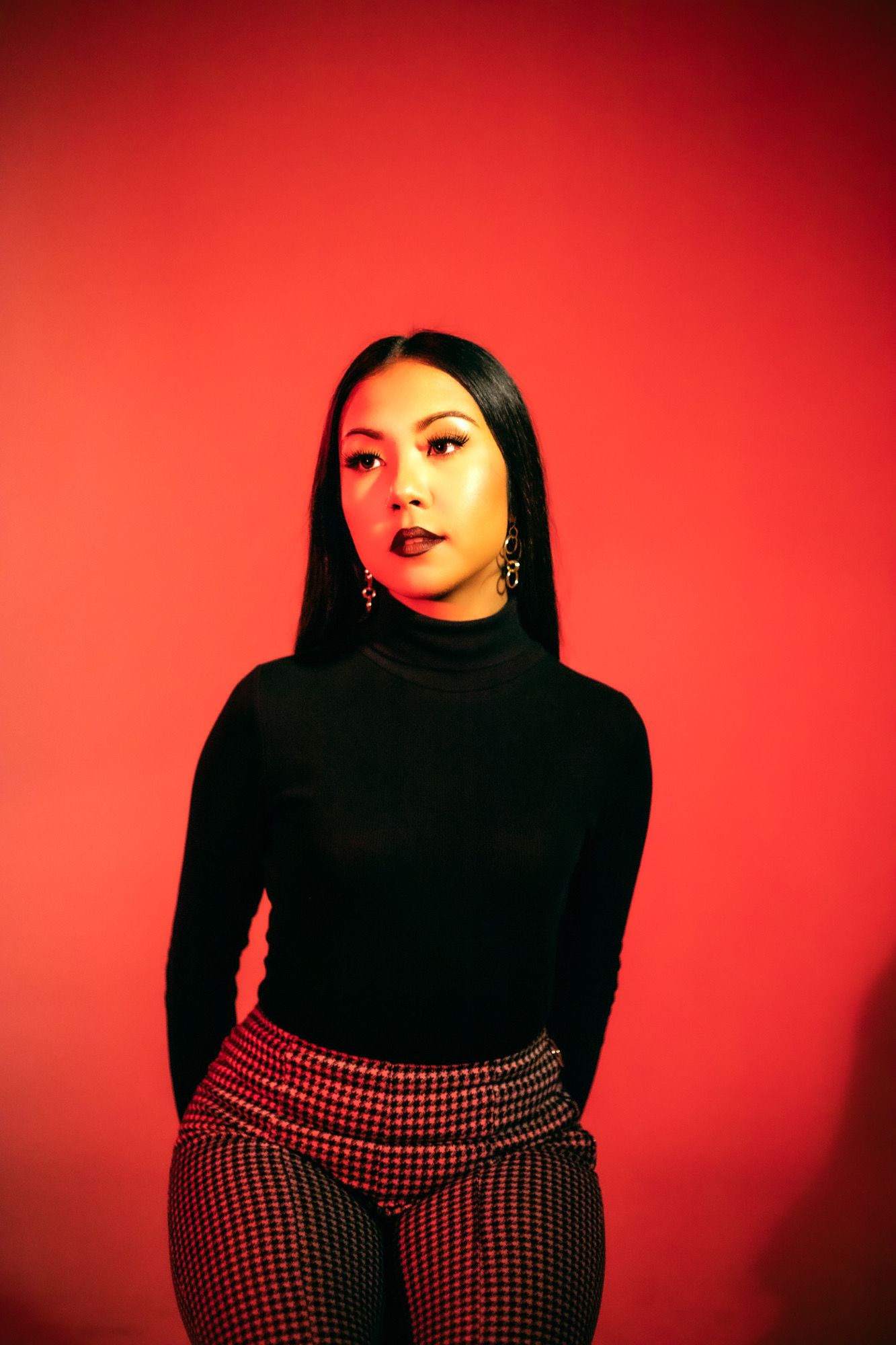 Young woman in black turtleneck and plaid pants poses confidently against a bold red background.