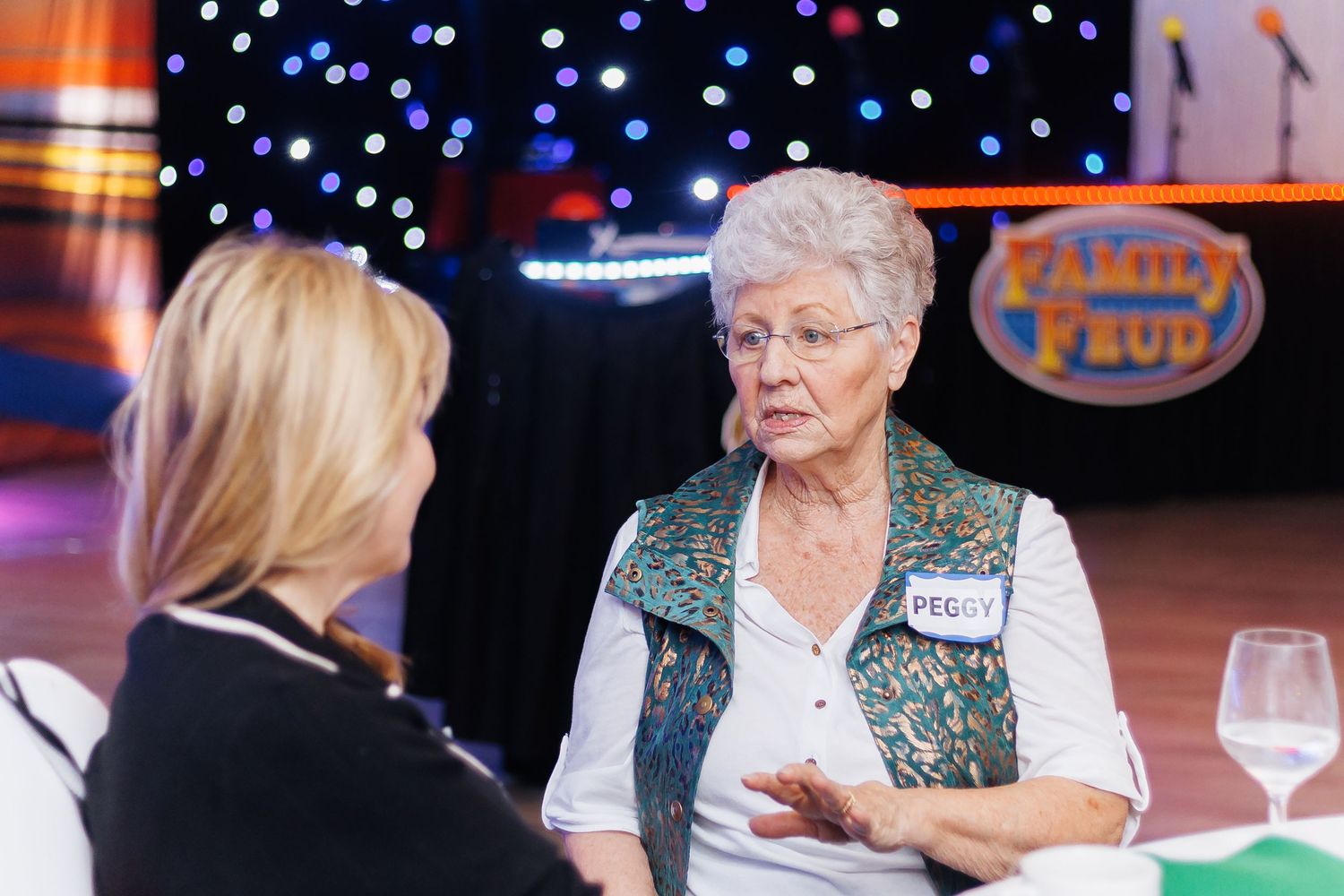 Elderly woman named Peggy conversing on Family Feud game show set with twinkling lights backdrop.