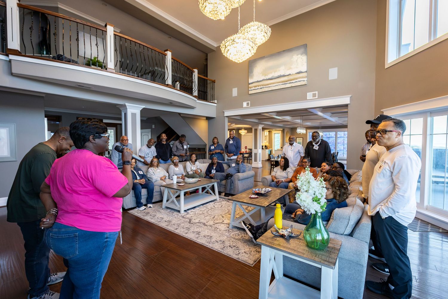 Group of people gathered in a spacious two-story home with hardwood floors and elegant chandelier lighting.
