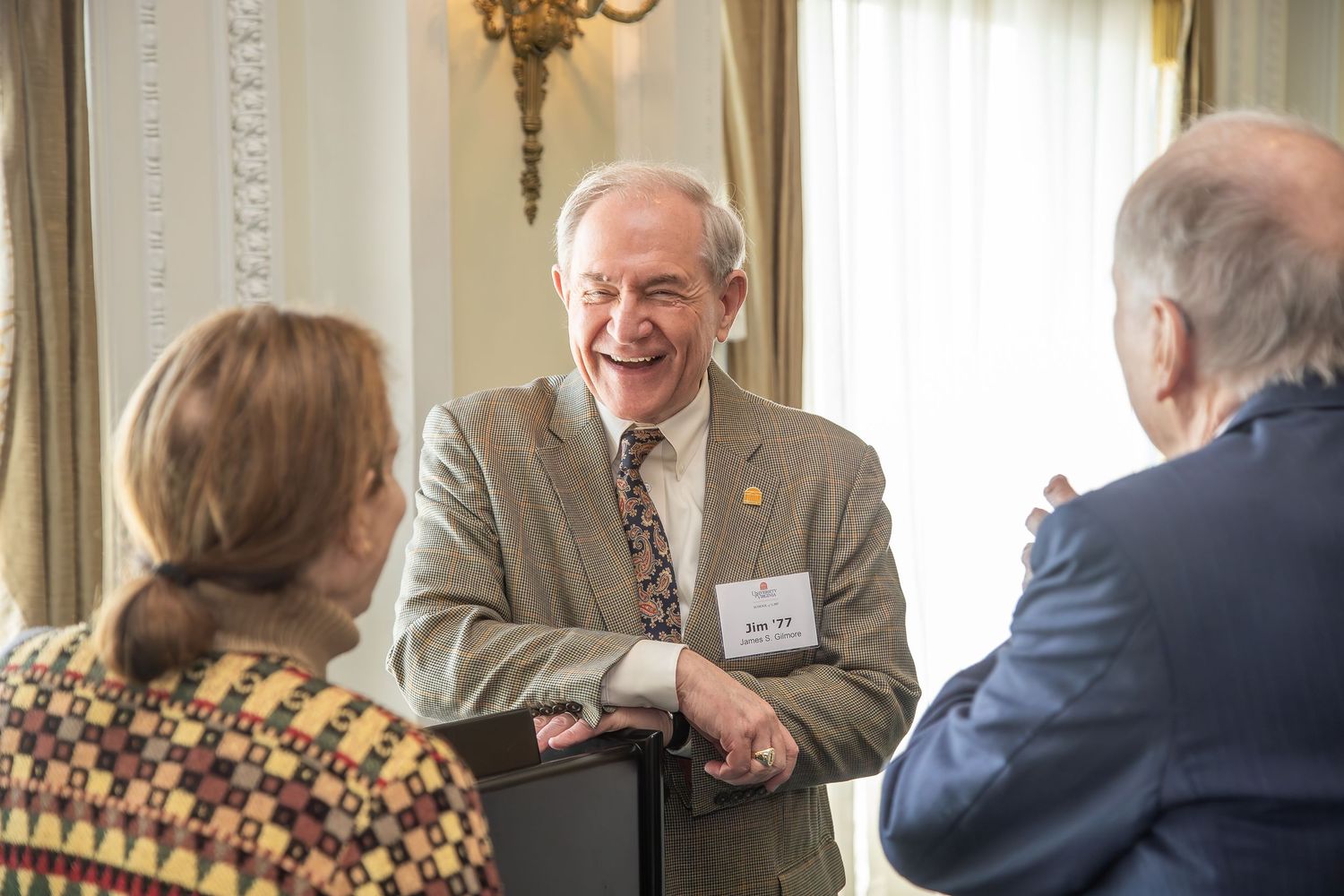 Elderly man in suit with name tag laughing joyfully while conversing with two people at a formal indoor event.