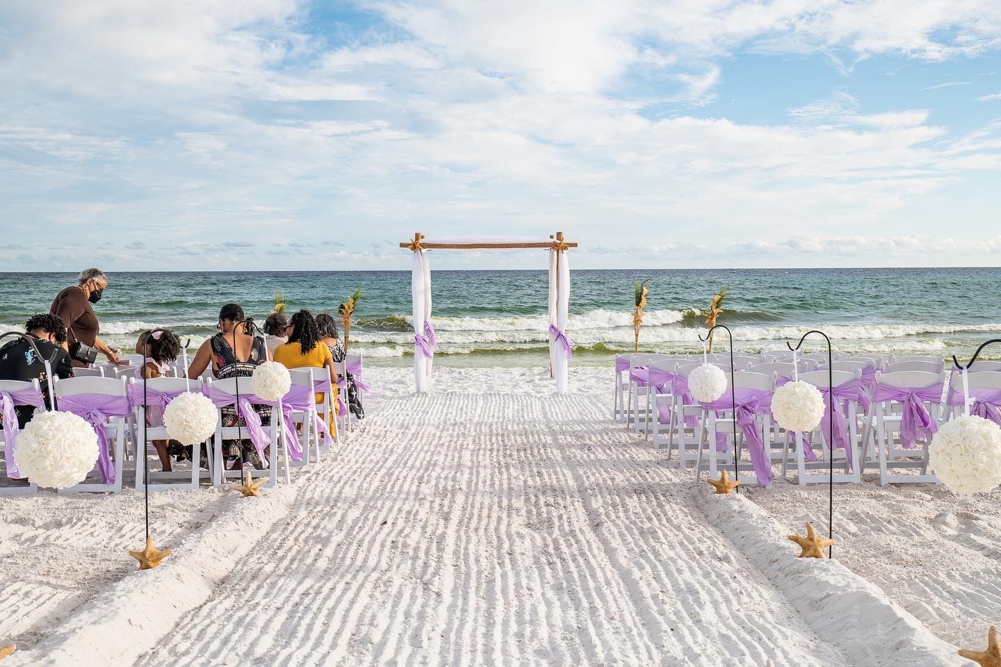Beach wedding setup with white chairs and purple accents along a sandy aisle.