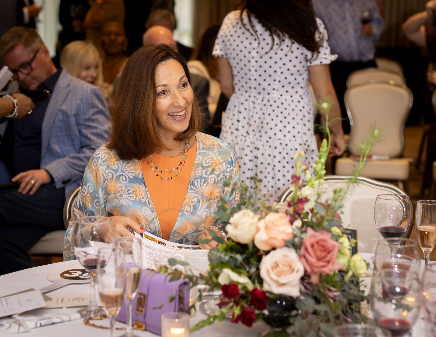 Woman in orange floral dress smiling at elegant event table with pink roses centerpiece and guests in background.