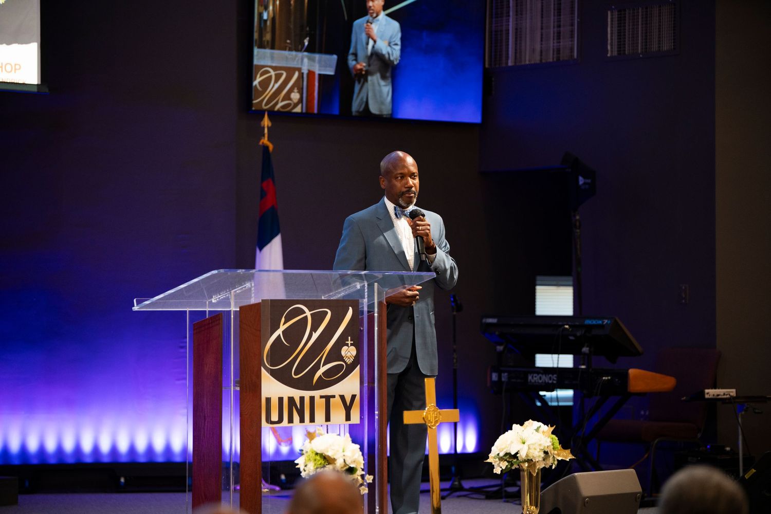 Man in suit speaking at Unity Church podium on stage with cross and floral arrangements.