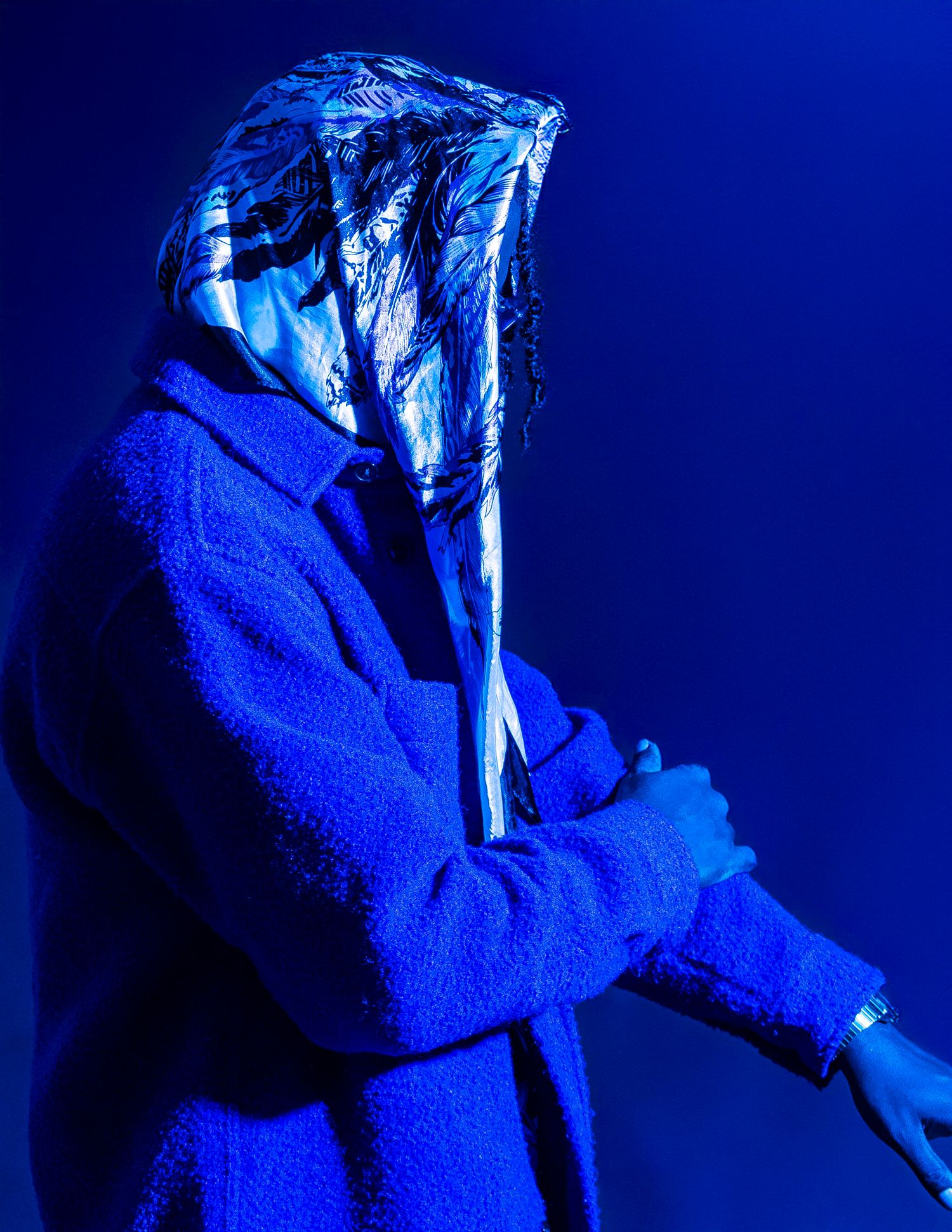 Person in blue coat with silver foil hood under dramatic blue lighting, arms crossed in artistic fashion pose.