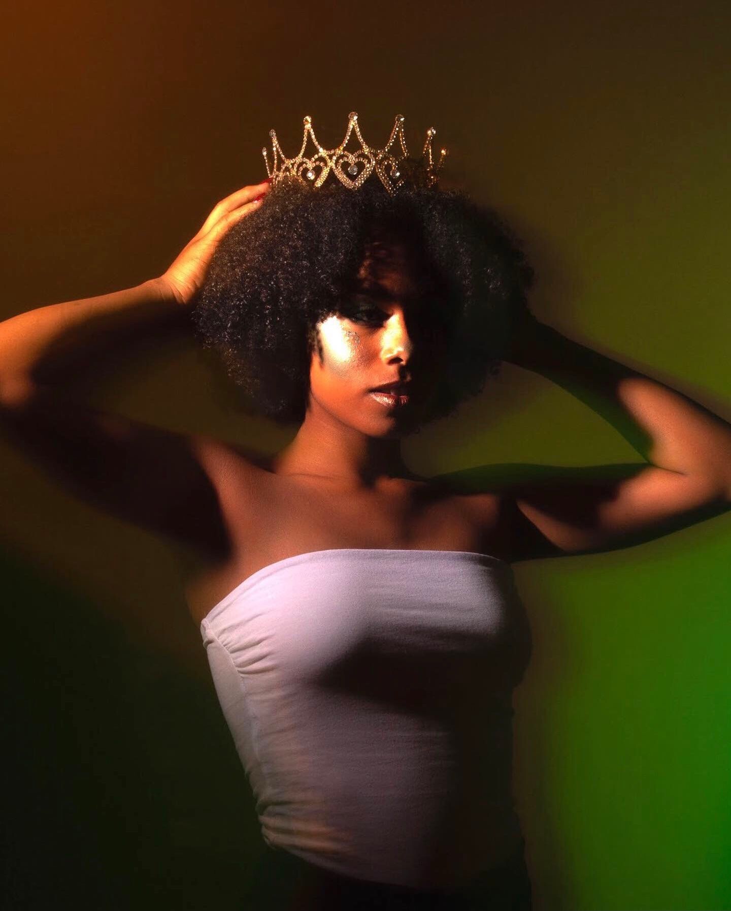 A Black woman with a natural afro wearing a white tube top and gold crown against a dramatic green and orange lit background.