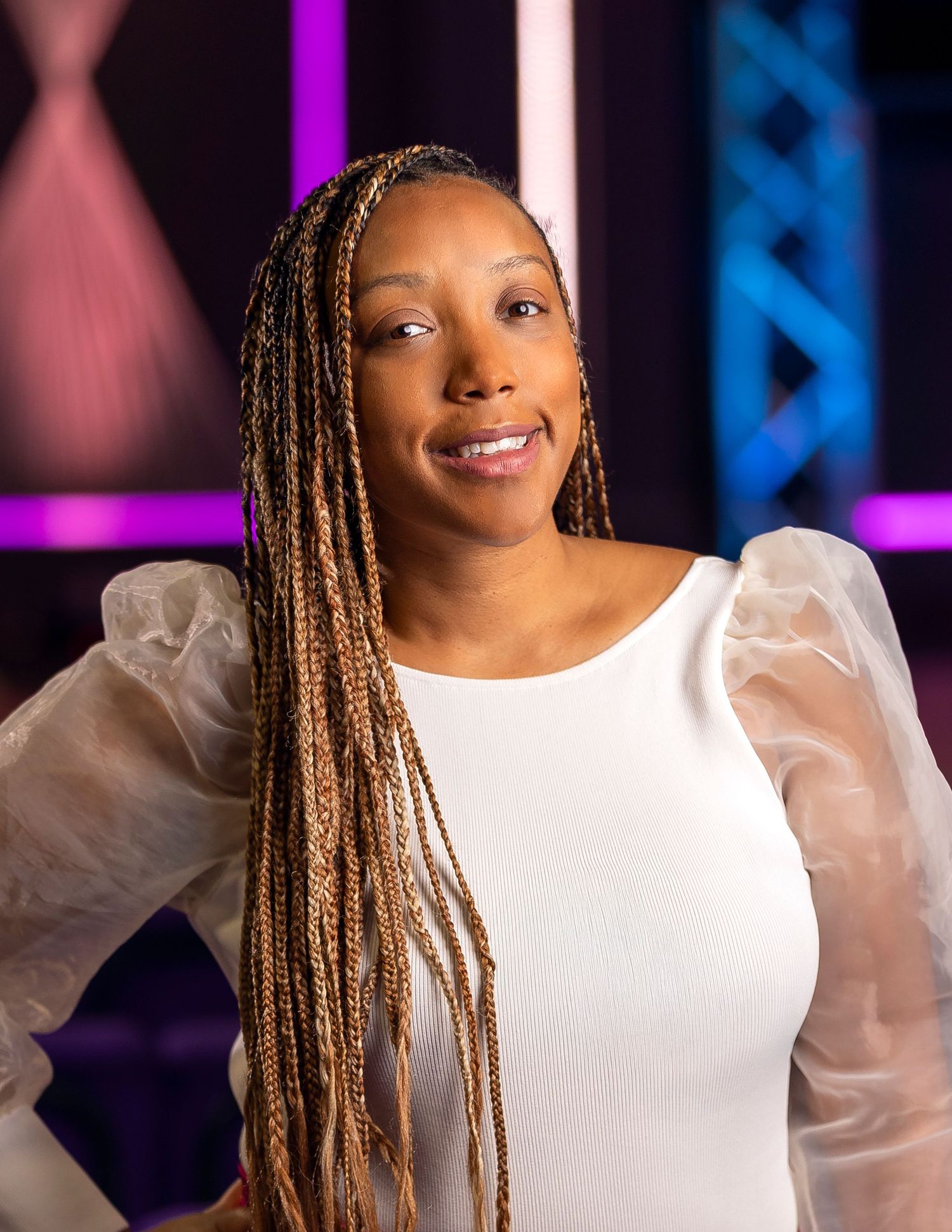 Woman with long box braids in white puff-sleeve top smiling confidently against colorful studio backdrop.