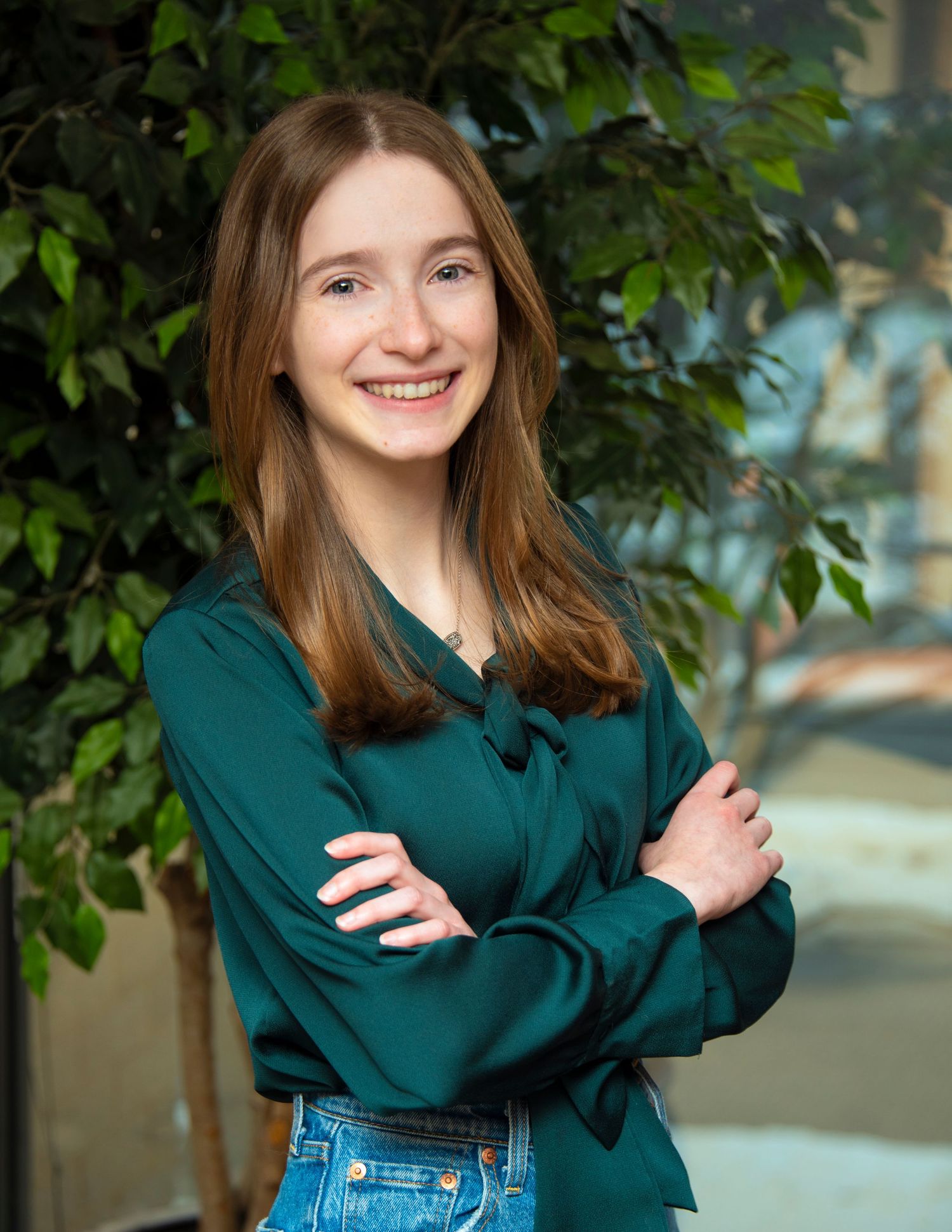 Young woman in teal blouse smiling with arms crossed, posing in front of green plants indoors.