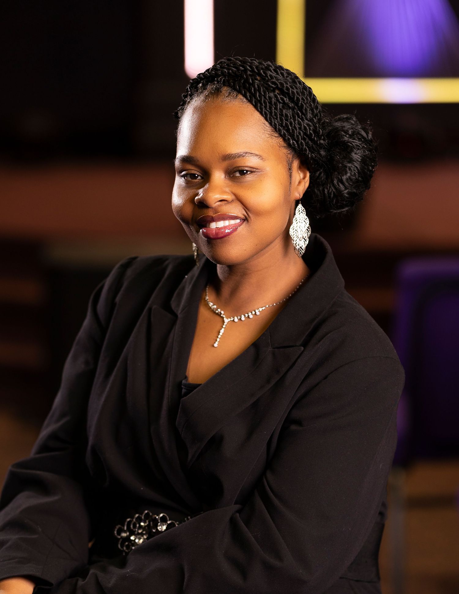 Smiling woman in black blazer with braided updo, wearing crystal earrings and necklace, seated in a professional studio setting.