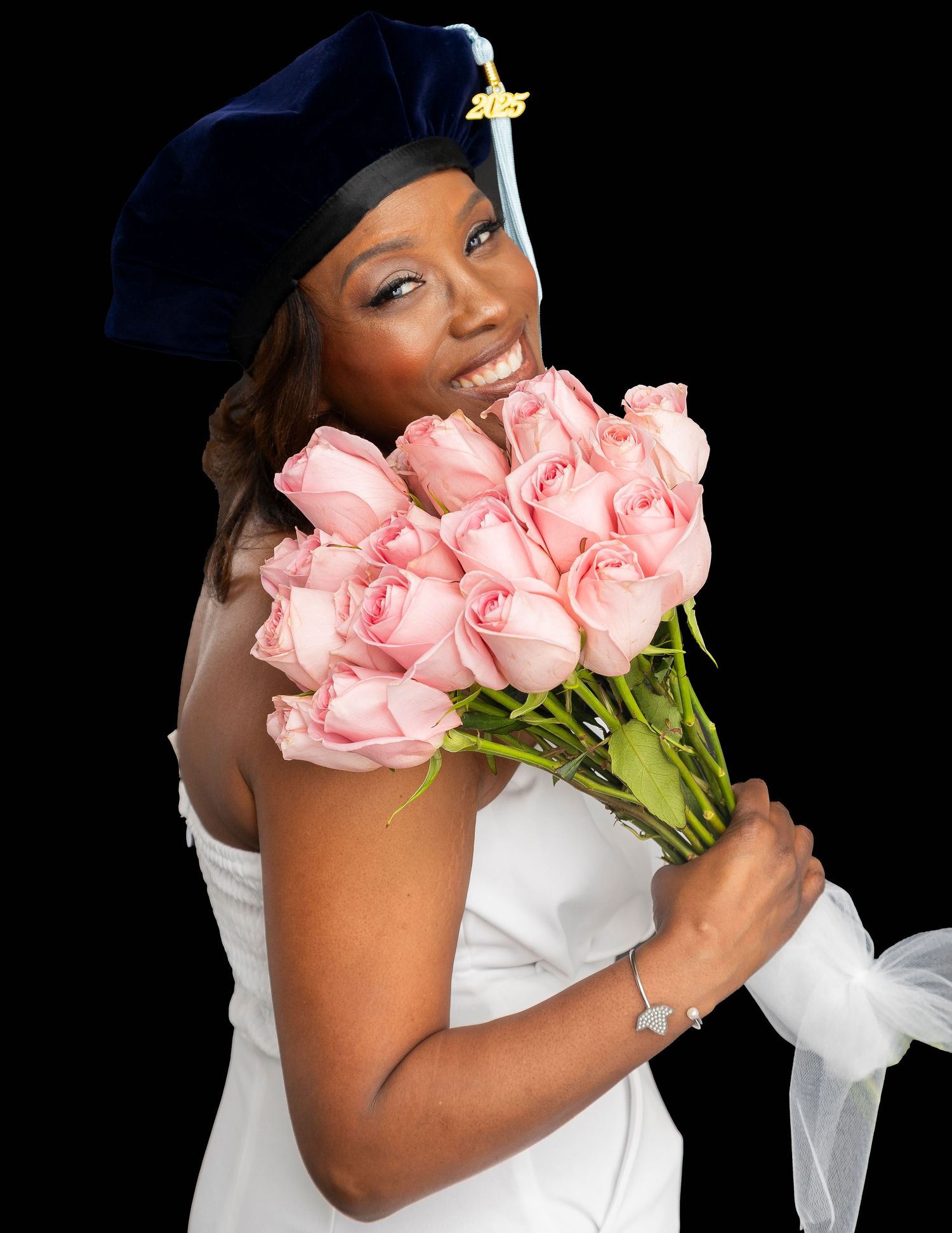 Smiling graduate in blue cap and white dress holding pink roses bouquet against black background.