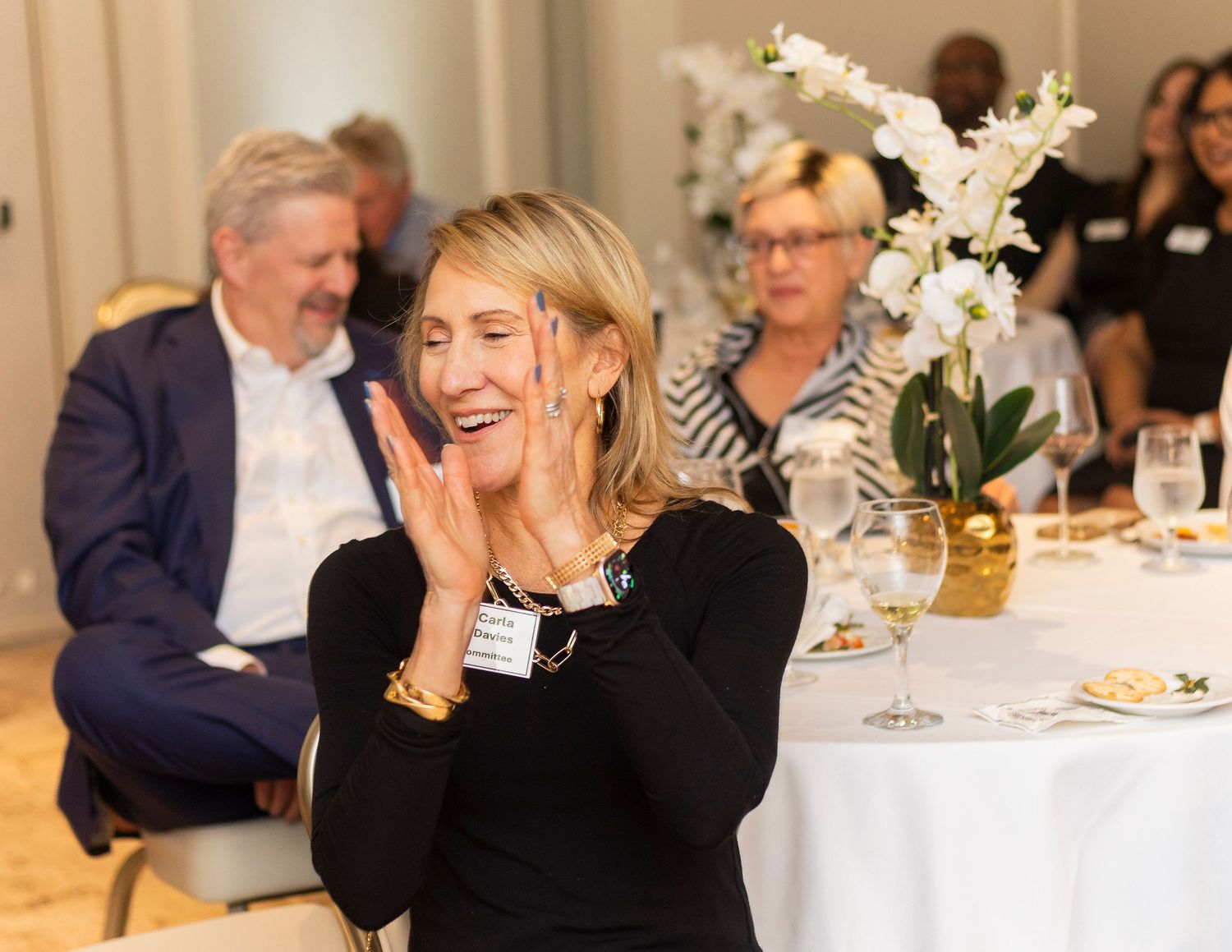Woman in black clapping at elegant dinner event with guests and white floral centerpiece in background.
