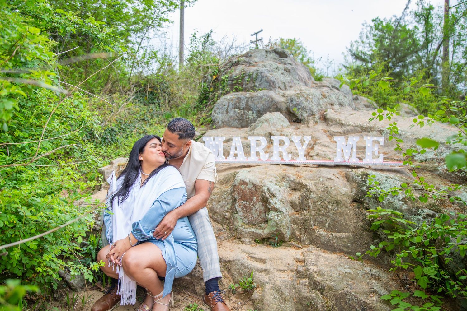 Couple posing near large rocks with 'Marry Me' sign visible in natural outdoor setting.