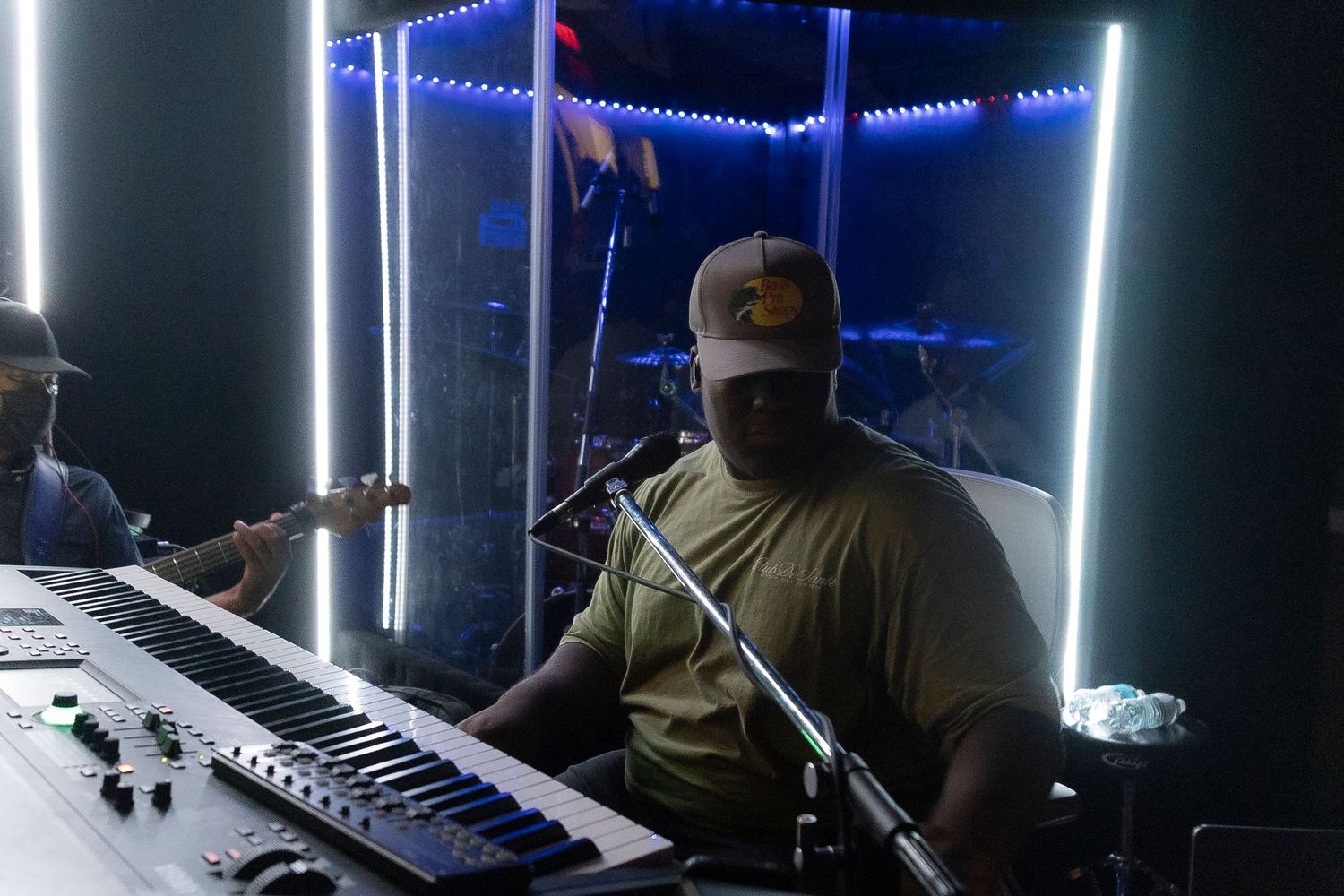 Two musicians perform in a neon-lit studio, one playing a keyboard synthesizer and another with a guitar in the background.