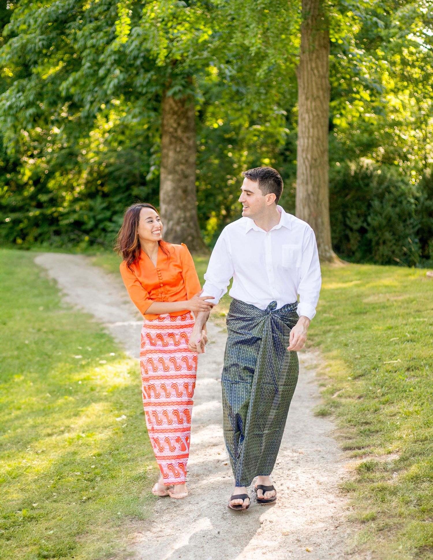 Couple walking on a wooded trail, wearing casual summer outfits in dappled sunlight.