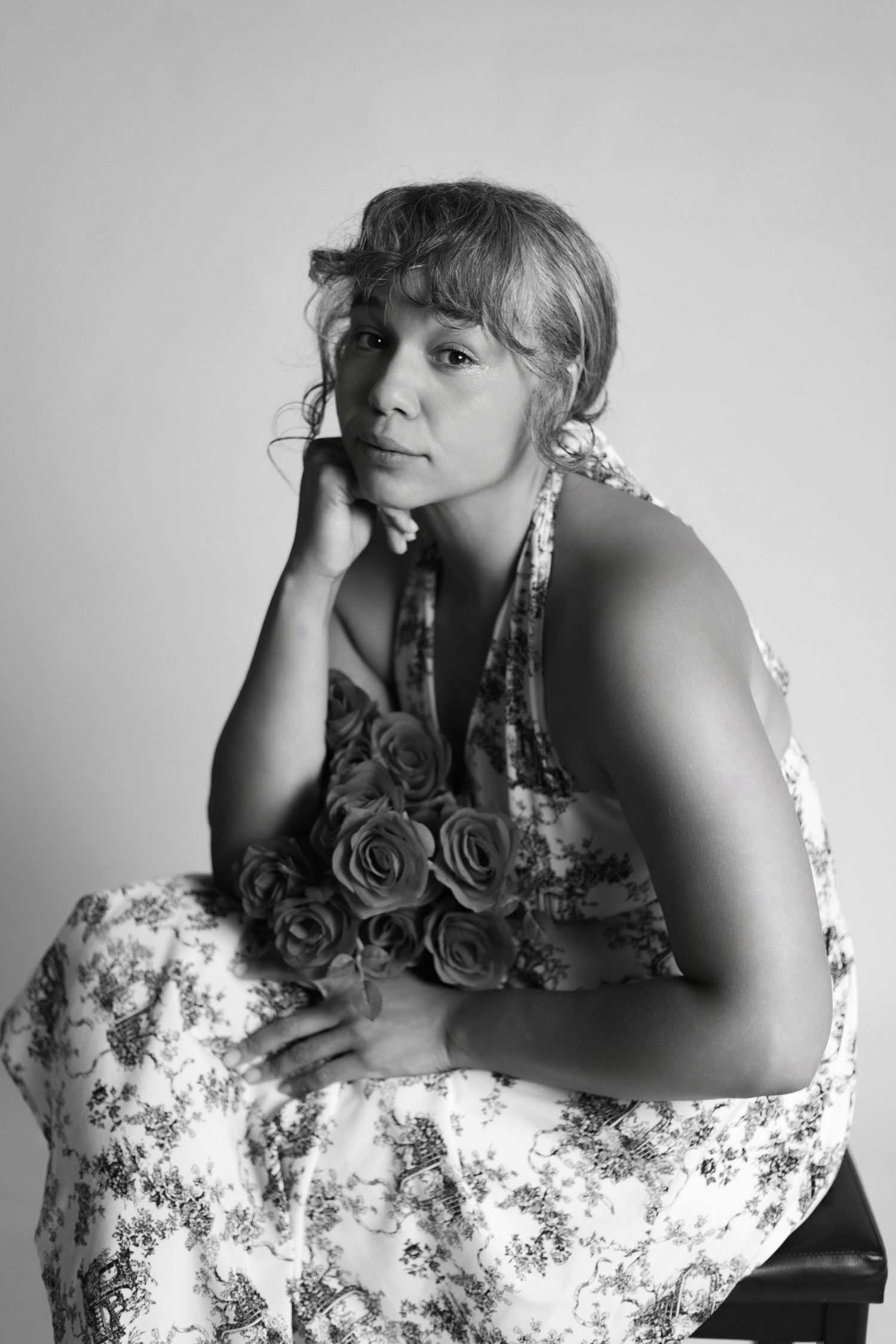 Black and white portrait of a woman in floral dress with rose necklace, resting chin on hand, seated pose.