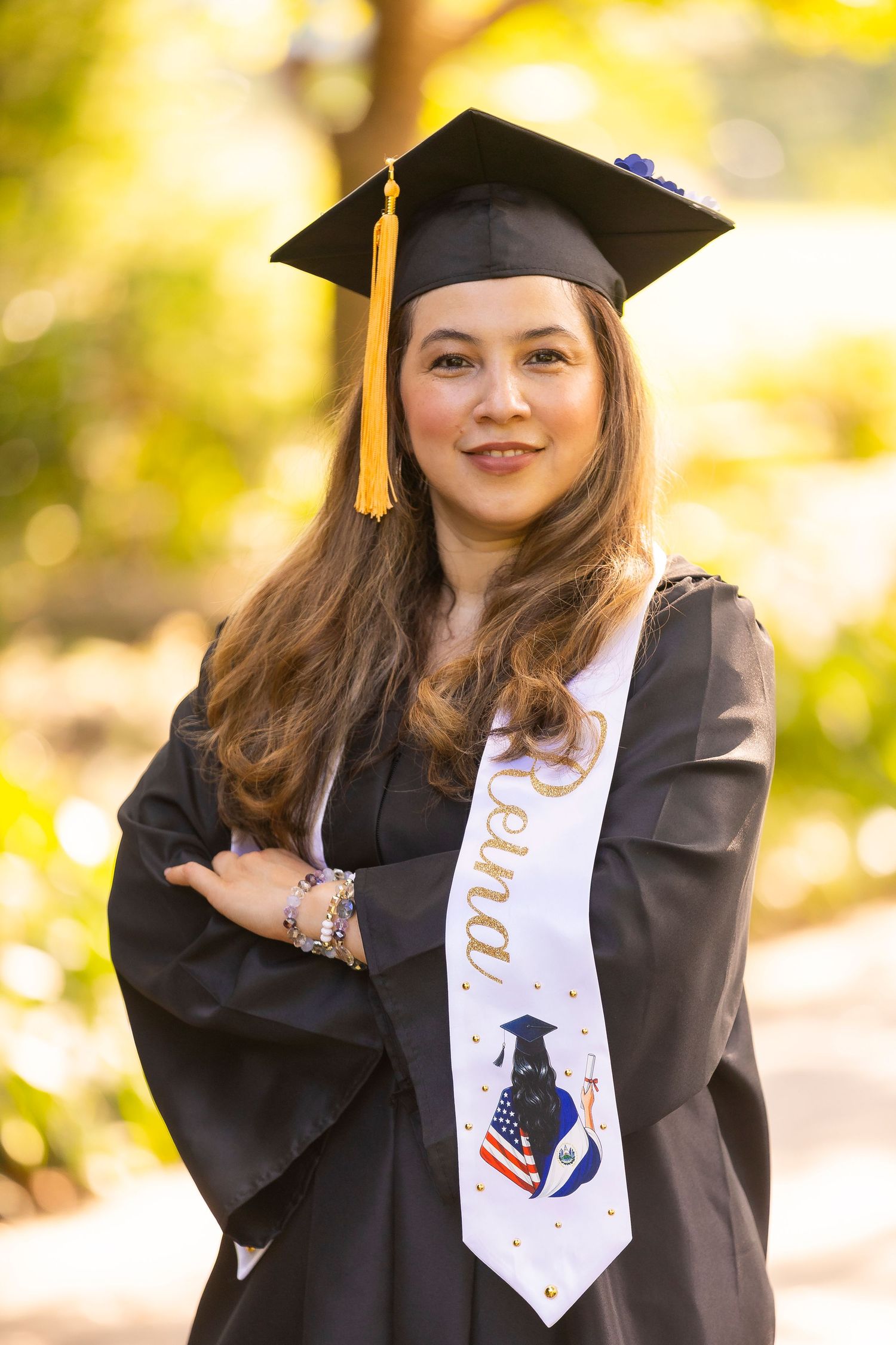 Young woman in black graduation cap and gown with a sash smiles outdoors with arms crossed.