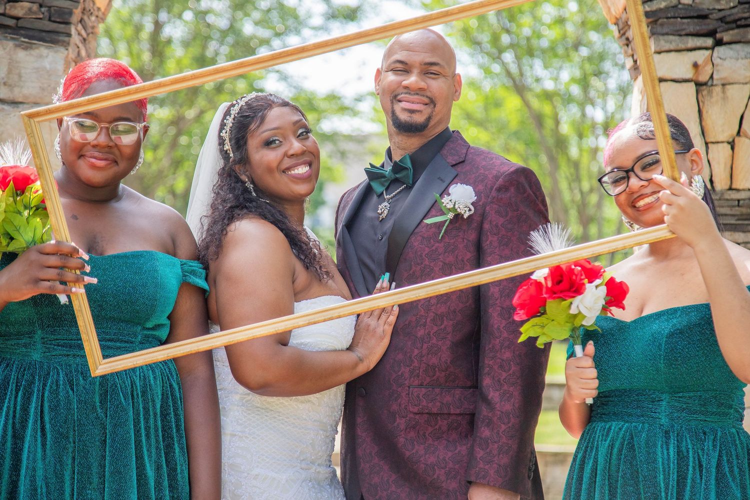 Wedding party members in teal dresses and burgundy suits posing with decorative frames outdoors.
