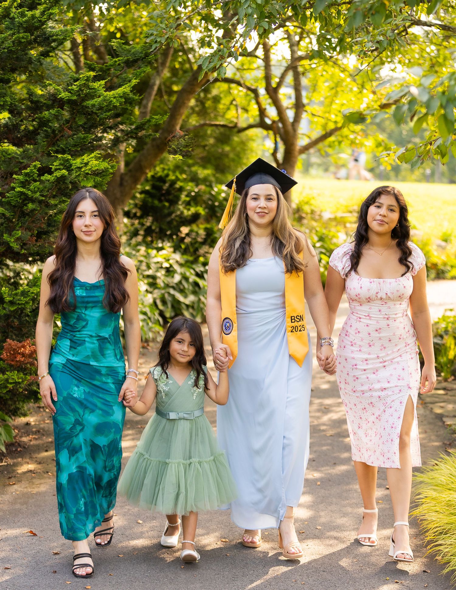 Four women walk together outdoors, one wearing a graduation cap and gold sash, celebrating a graduation milestone.