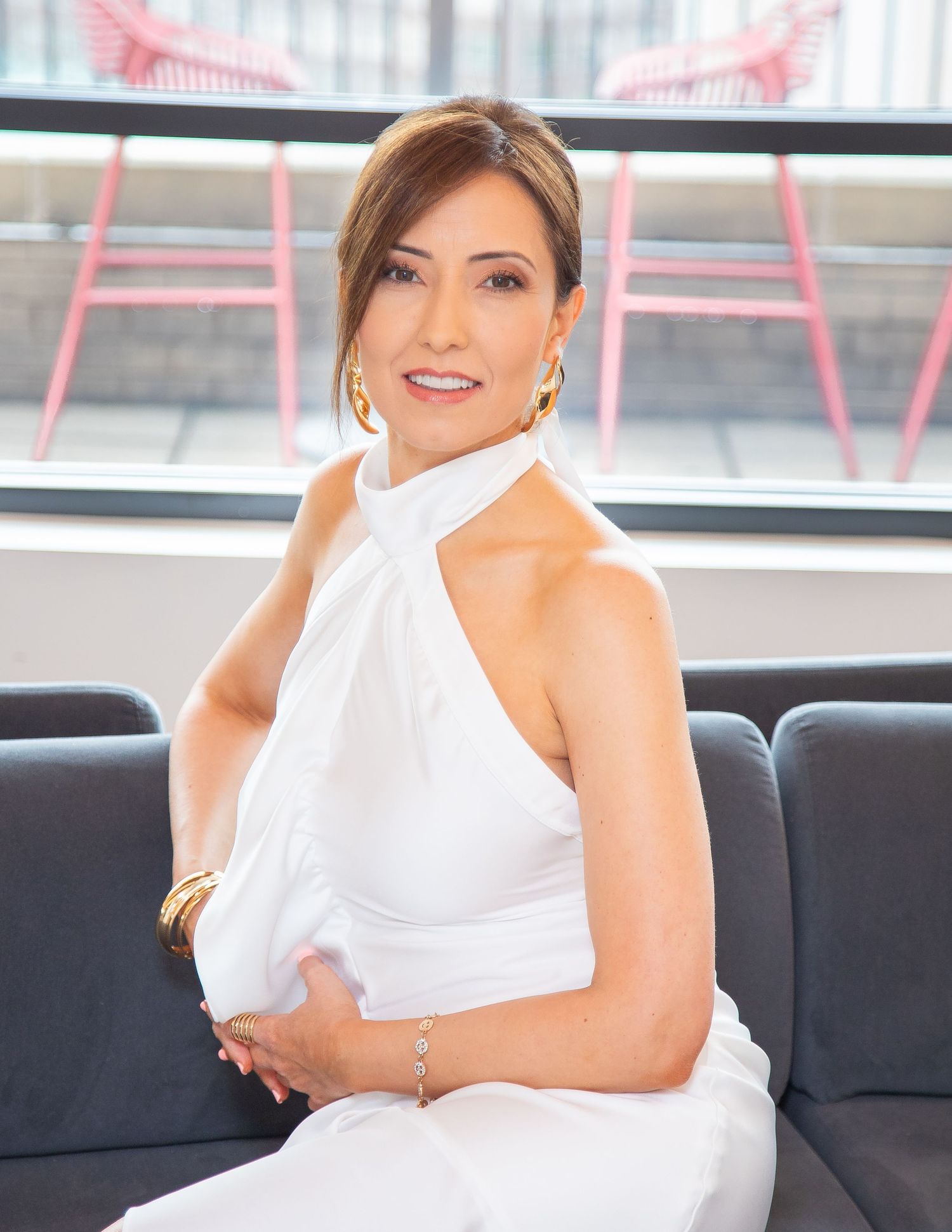 Elegant woman in white halter dress seated on gray sofa, wearing gold jewelry, smiling confidently indoors.