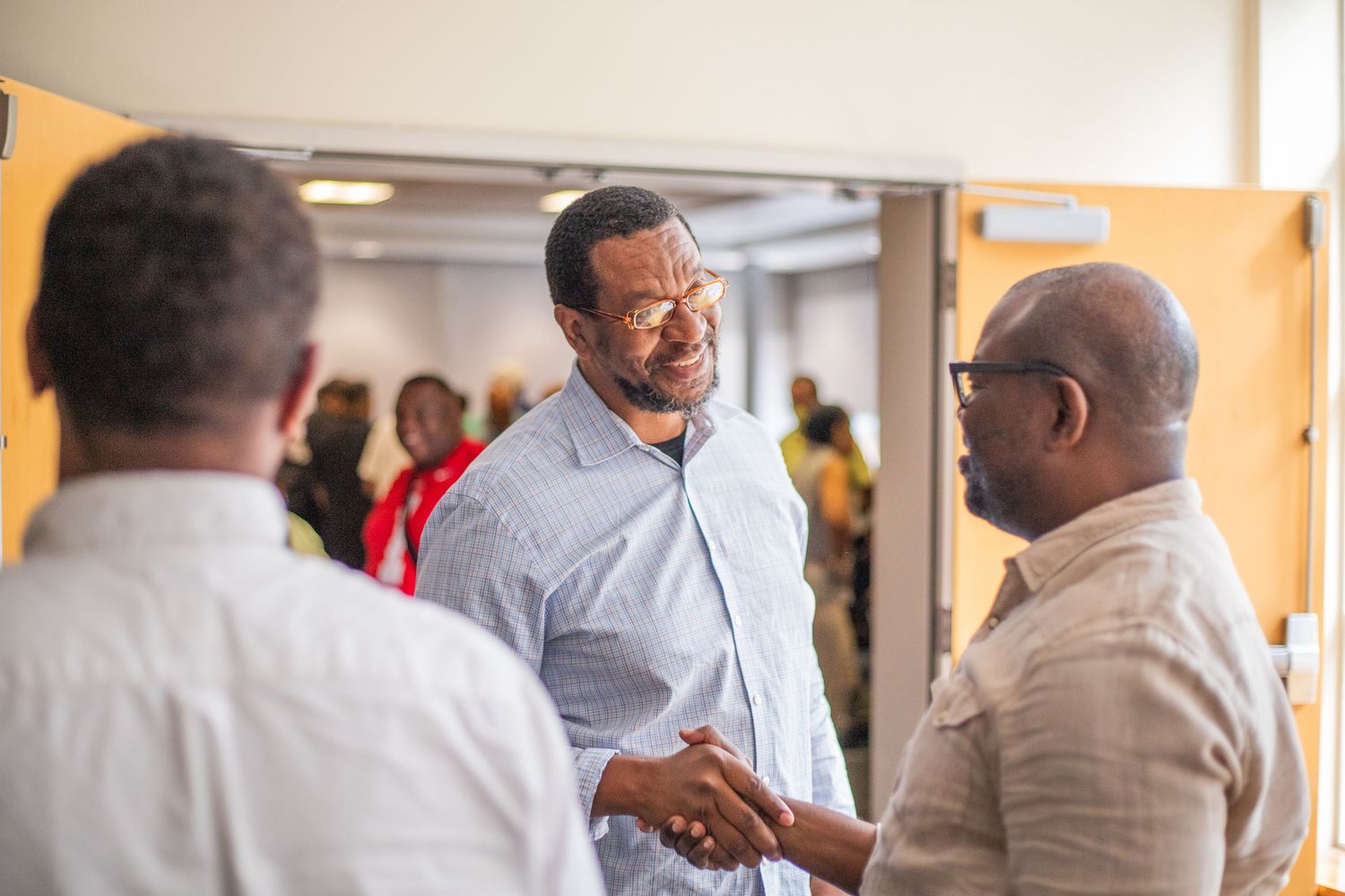 Men shaking hands and networking at an indoor professional event with diverse attendees in the background.