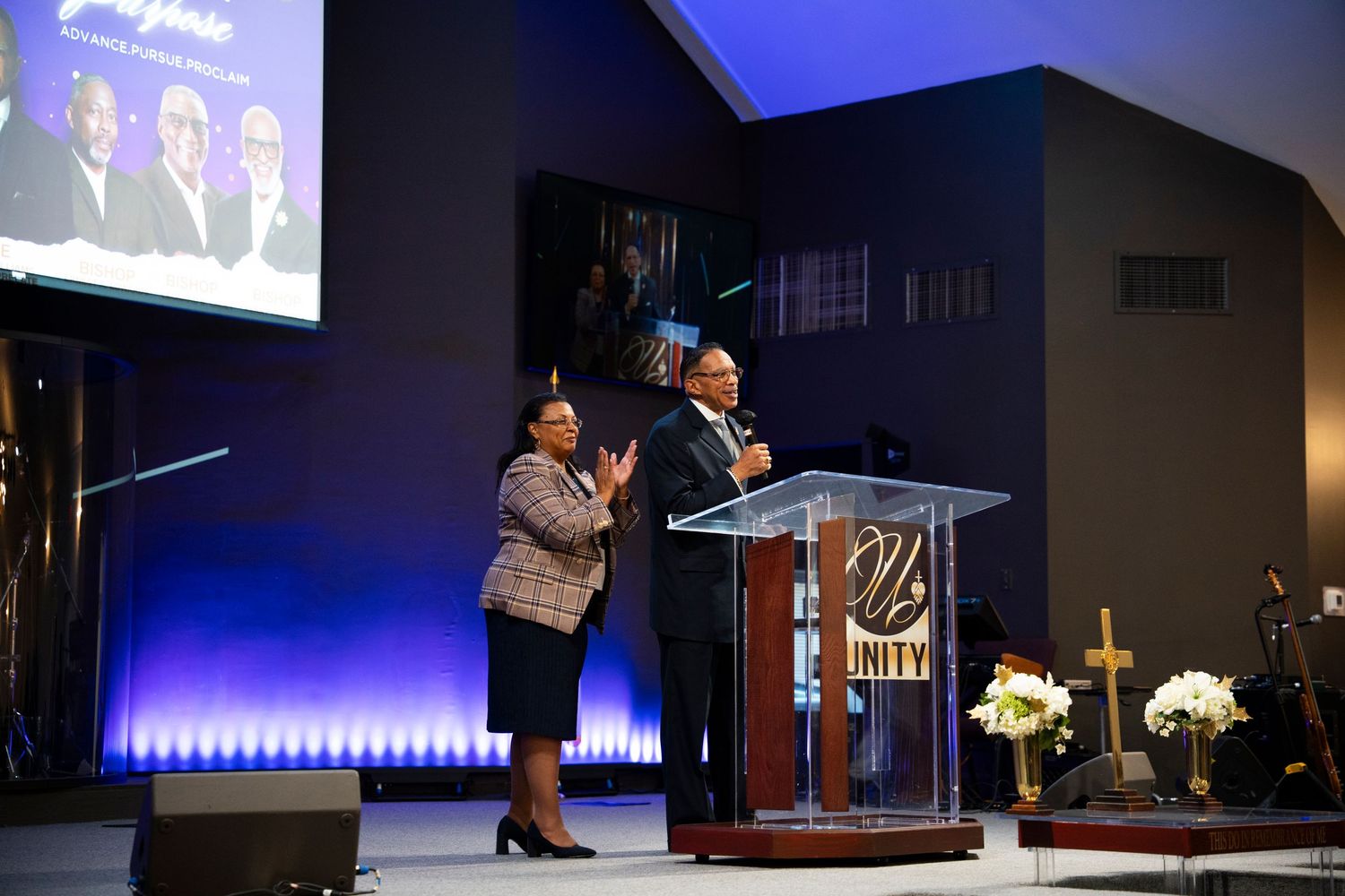 Two speakers on stage at a church podium with blue lighting, a cross, and a projection screen behind them.