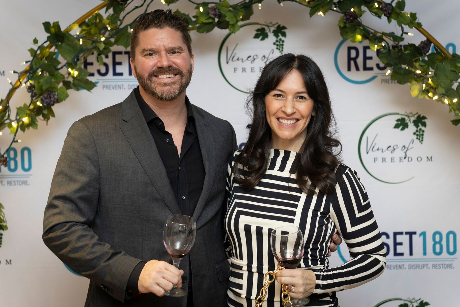 A man and woman smile holding wine glasses at a Reset180 Vineyard Premium event backdrop.