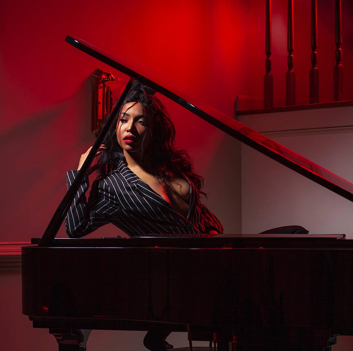 Woman in striped blazer poses elegantly at a black grand piano bathed in dramatic red light.