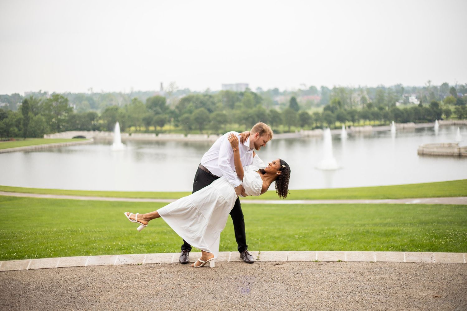 Couple performing romantic dip pose by lakeside with fountains in background.