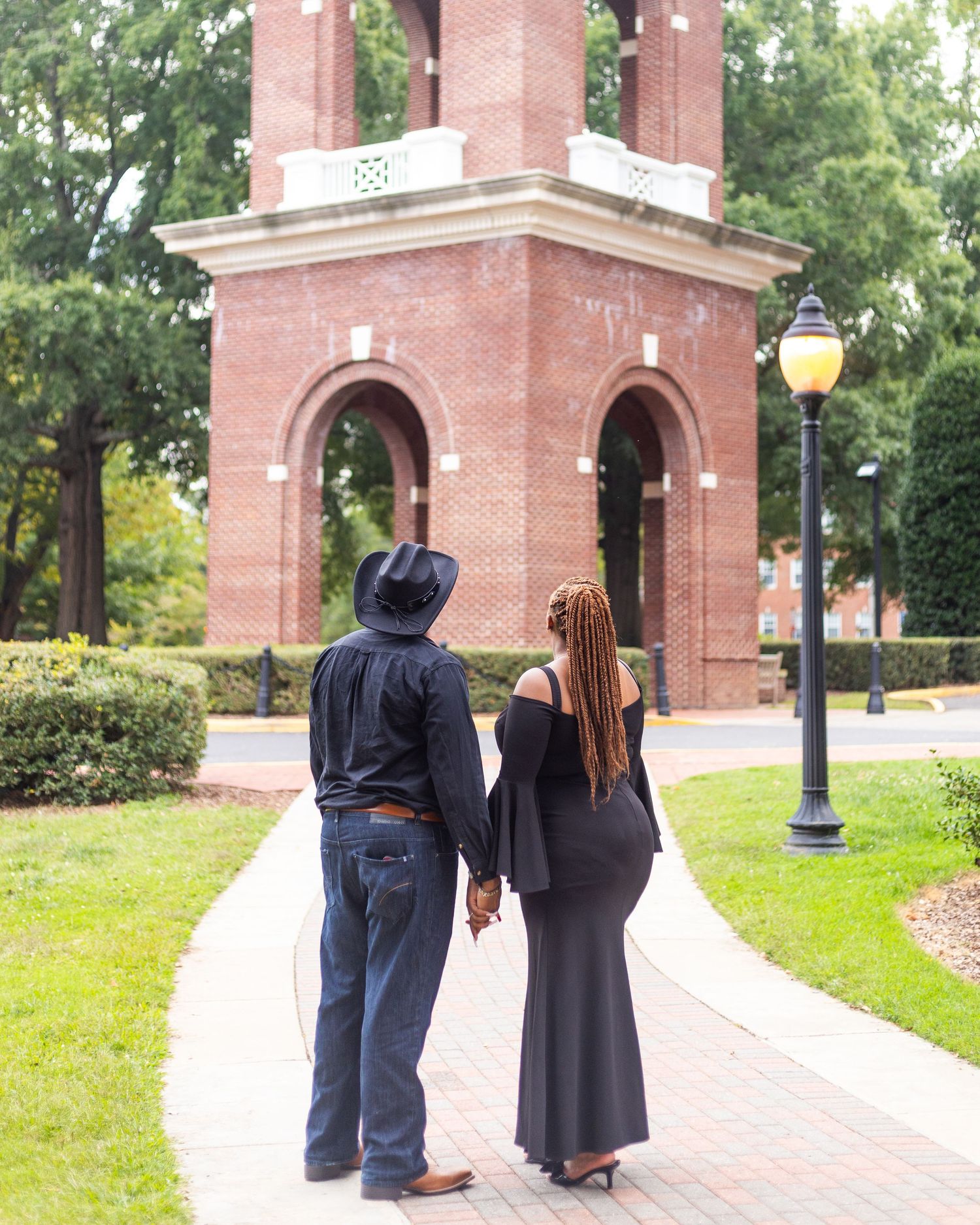 A couple walking together on a brick pathway near red brick archways and green landscaping.
