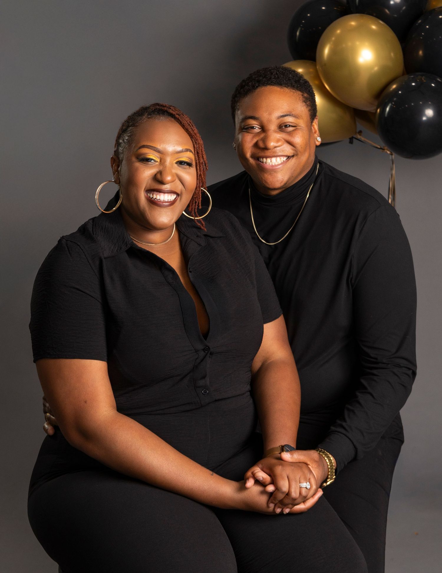 Two women in black outfits smile together in front of gold and black balloons in a studio portrait.